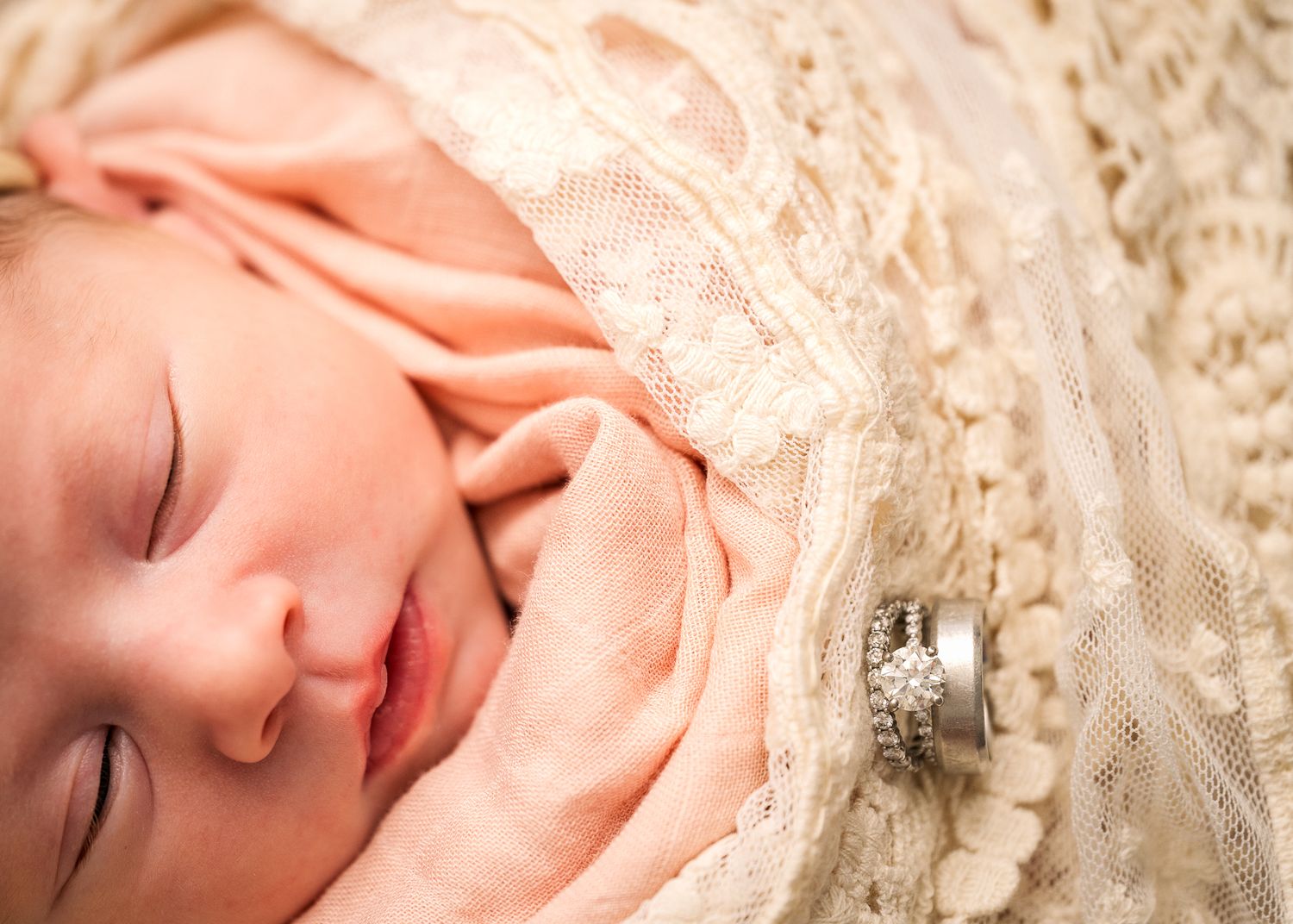 Sleeping newborn wrapped in peach blanket against white lace background.