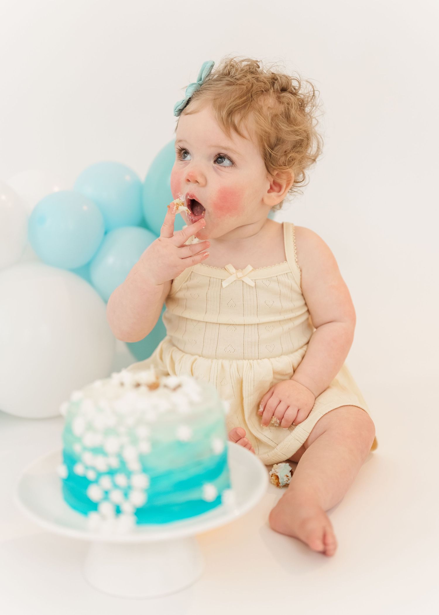 Cake smash. Baby in cream dress smiles while reaching for white and turquoise birthday cake during first birthday cake smash photoshoot.