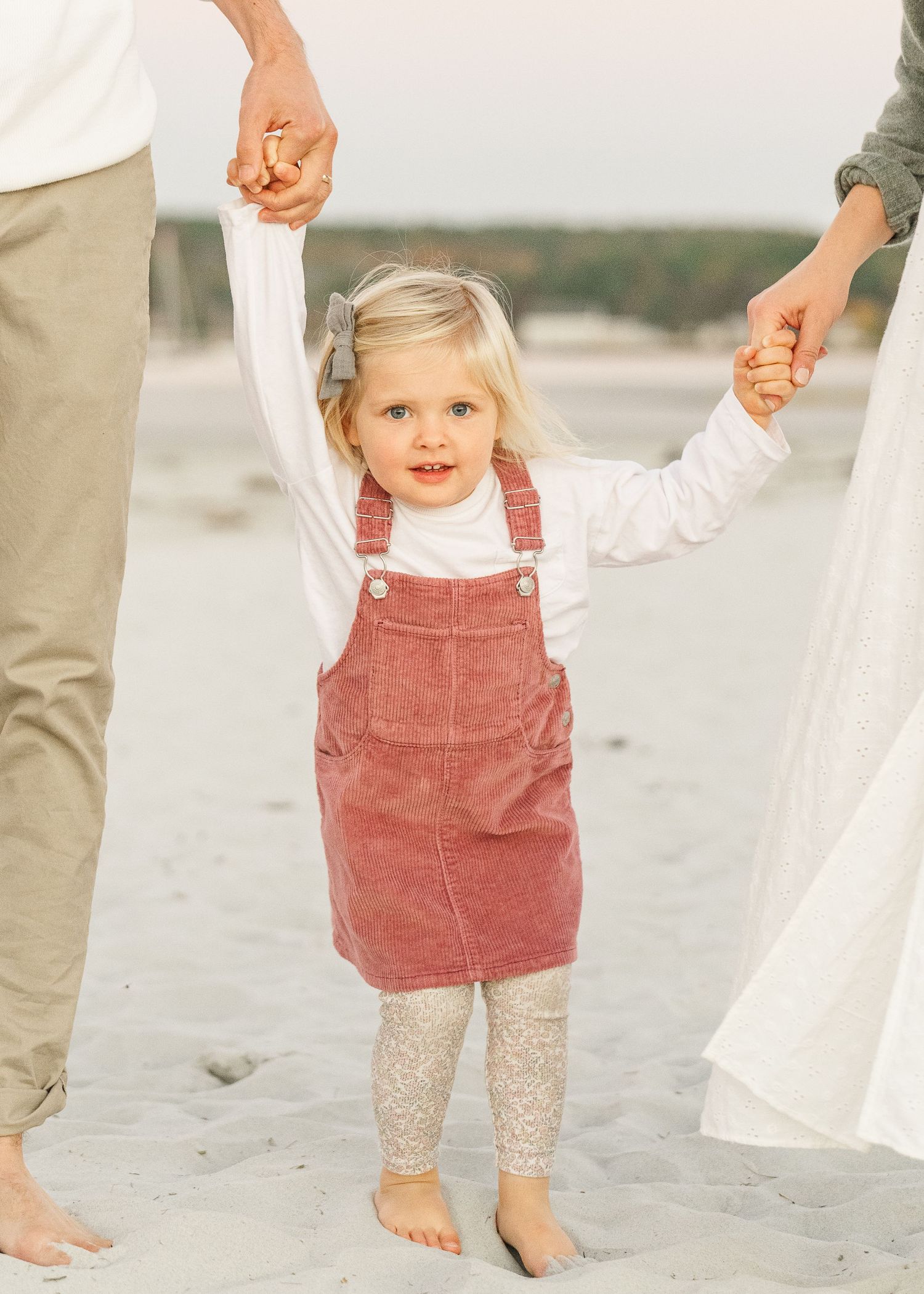 Small child wearing red corduroy dress and sparkly tights walks barefoot on beach sand holding hands with adults.