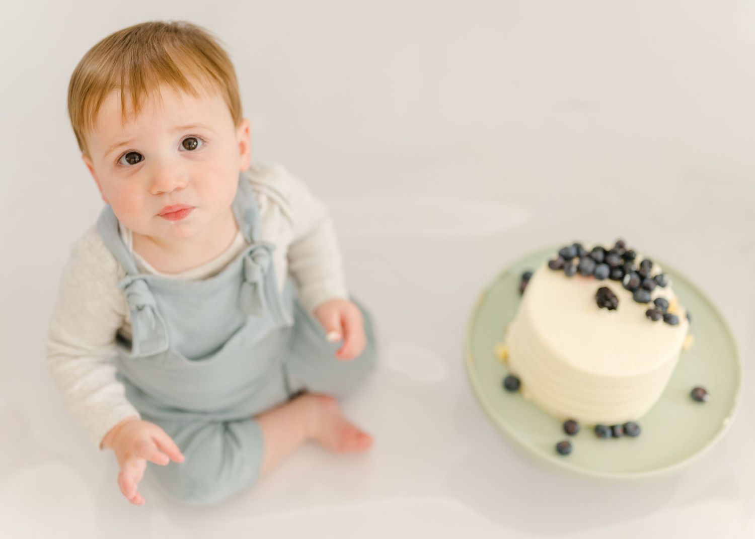 Baby in light blue overalls sits next to a white cake decorated with blueberries on a plate.