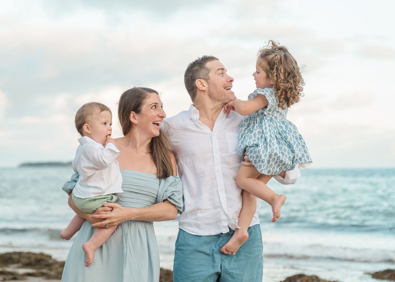 A cheerful family laughs together while standing on a rocky beach at sunset with ocean waves in the background.