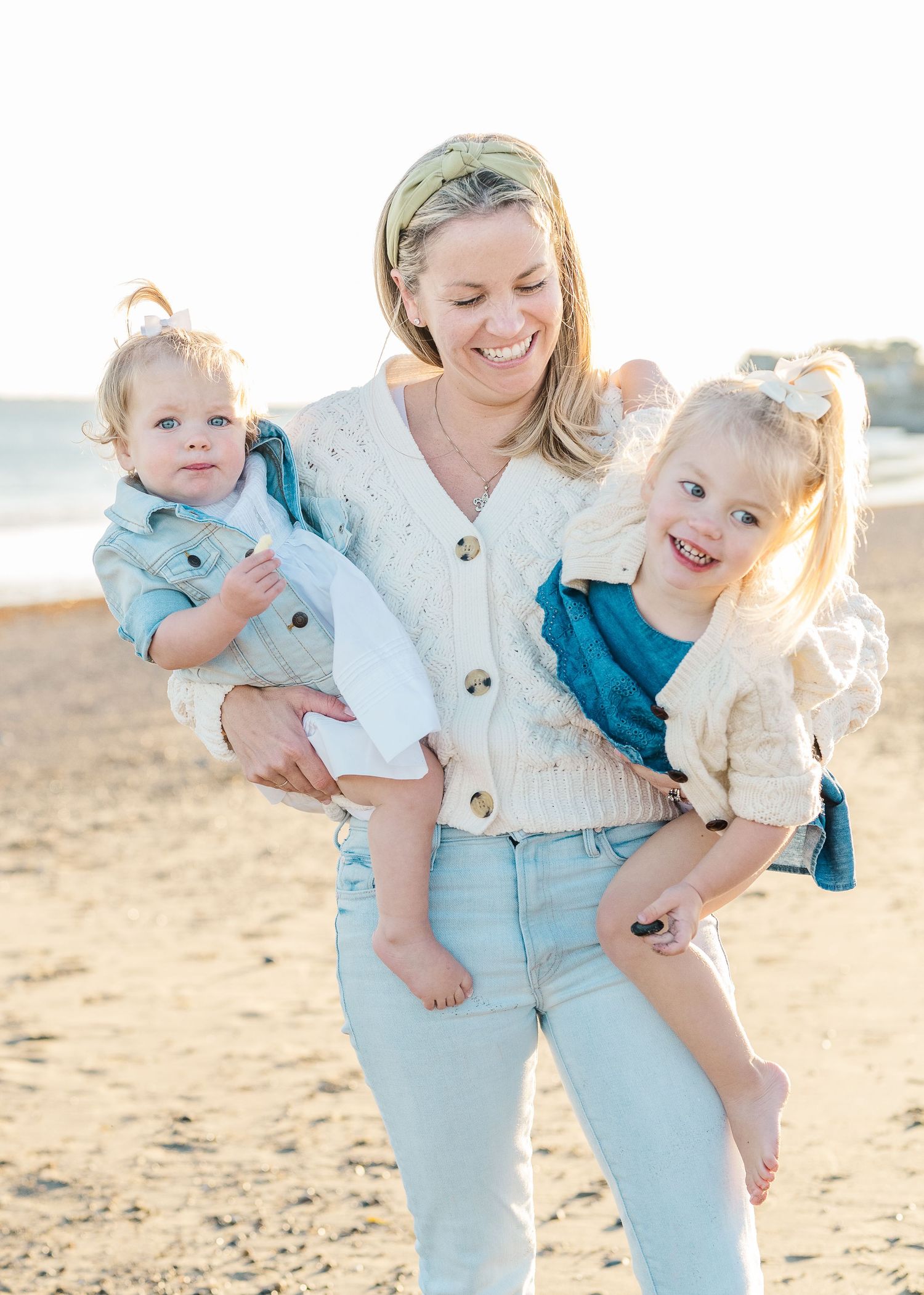 Mother in white cardigan holding two children on a sunlit beach.