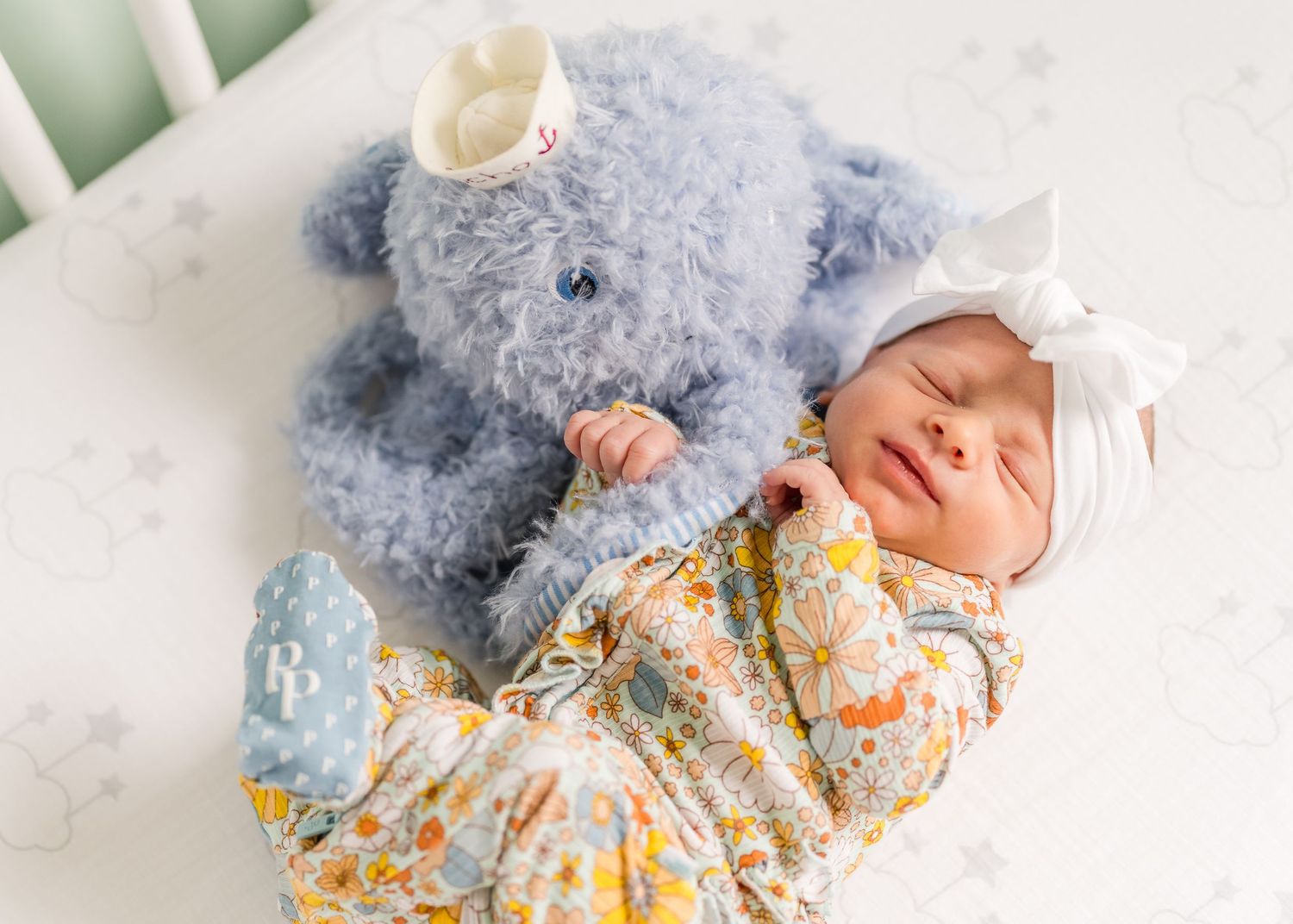 A sleeping newborn in floral dress next to blue plush elephant toy in crib.