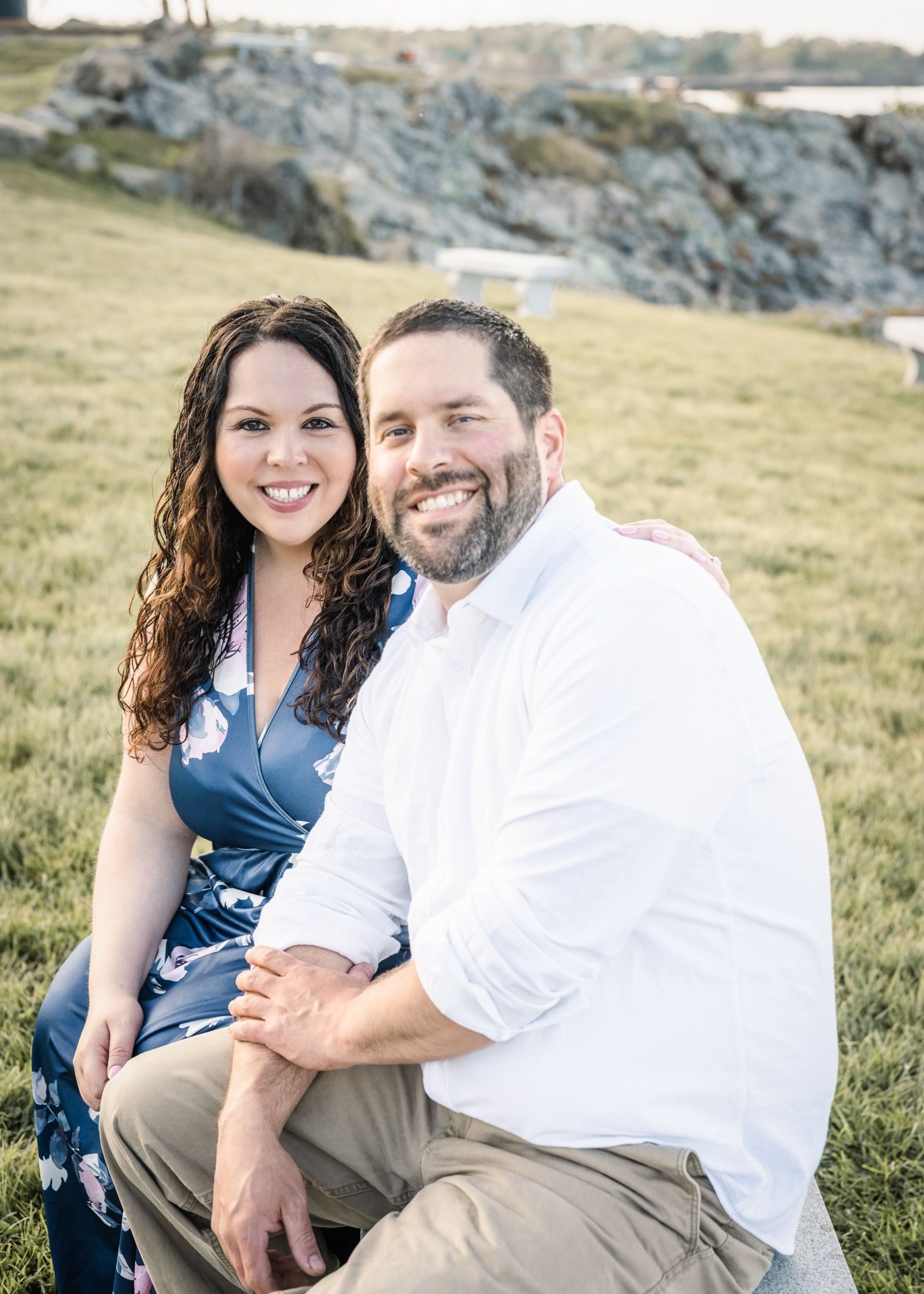 Happy couple sits together on grassy hill with rocky coastline in background.