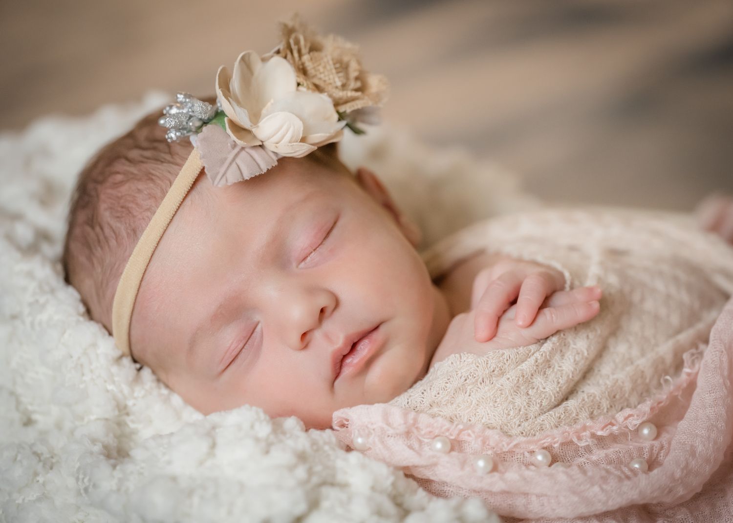 Peaceful sleeping newborn wearing delicate floral headband on cream textured blanket.