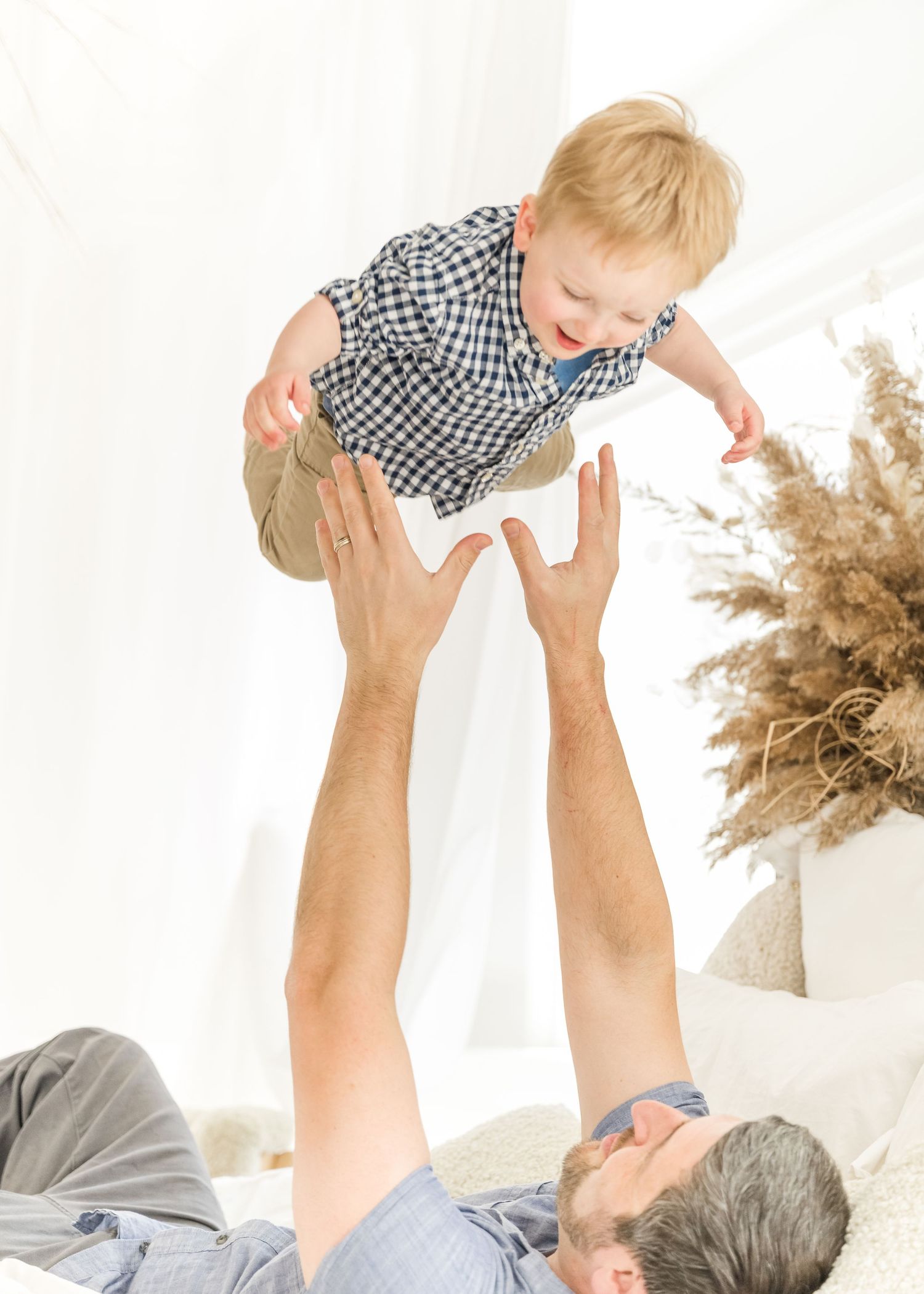 A joyful moment between a child in a checkered shirt being lifted up by adult hands with dried pampas grass decor nearby.