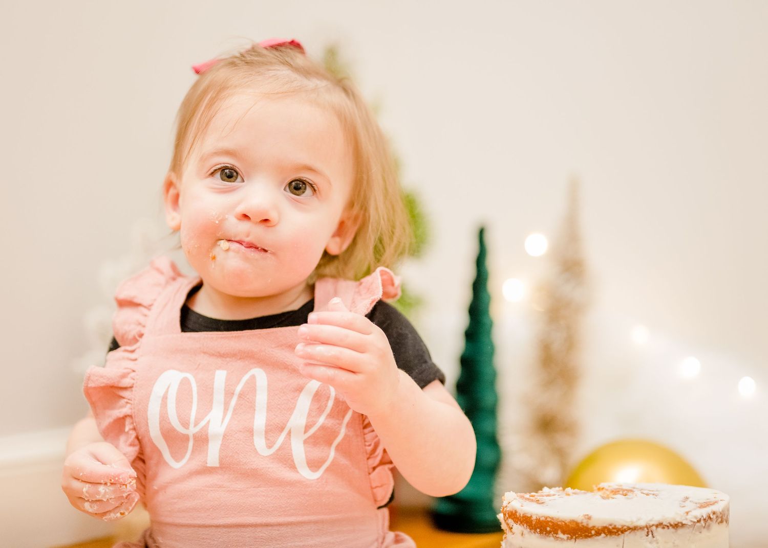 A toddler wearing a pink 'ONE' shirt smiles during birthday celebrations.