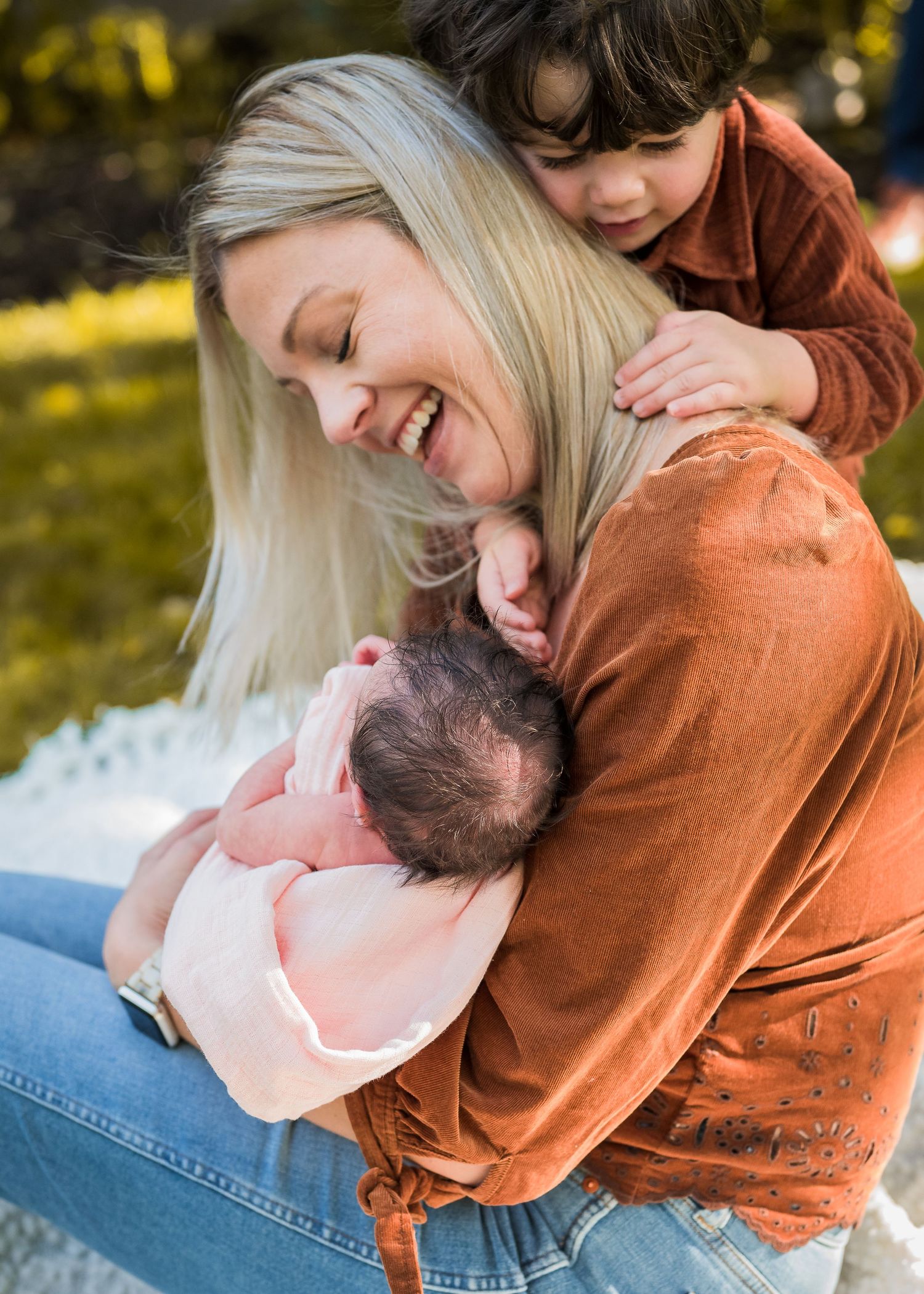 Joyful family moment outdoors with mother holding baby while sibling plays.
