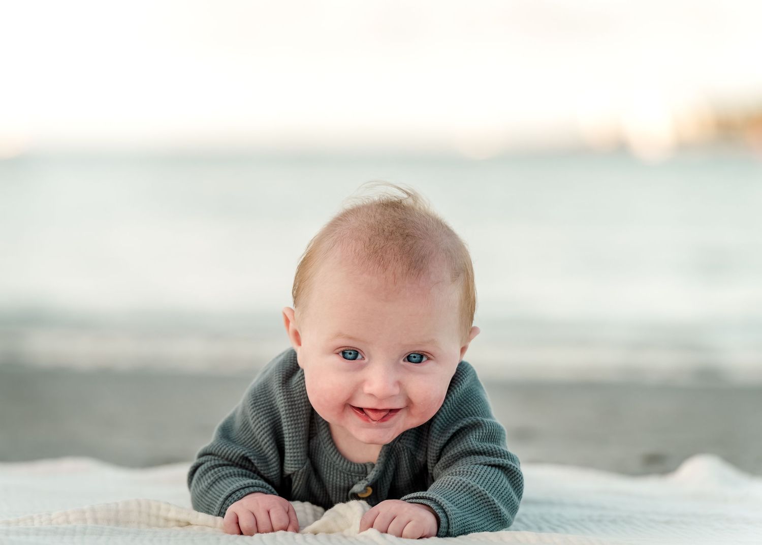 A smiling baby wearing a green sweater lies on a blanket at the beach during sunset.