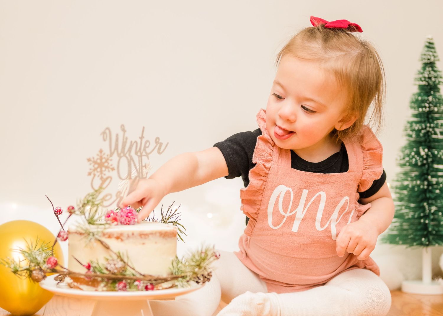 Small child wearing a pink 'one' bib explores a cake and decorations during a festive holiday photo session.