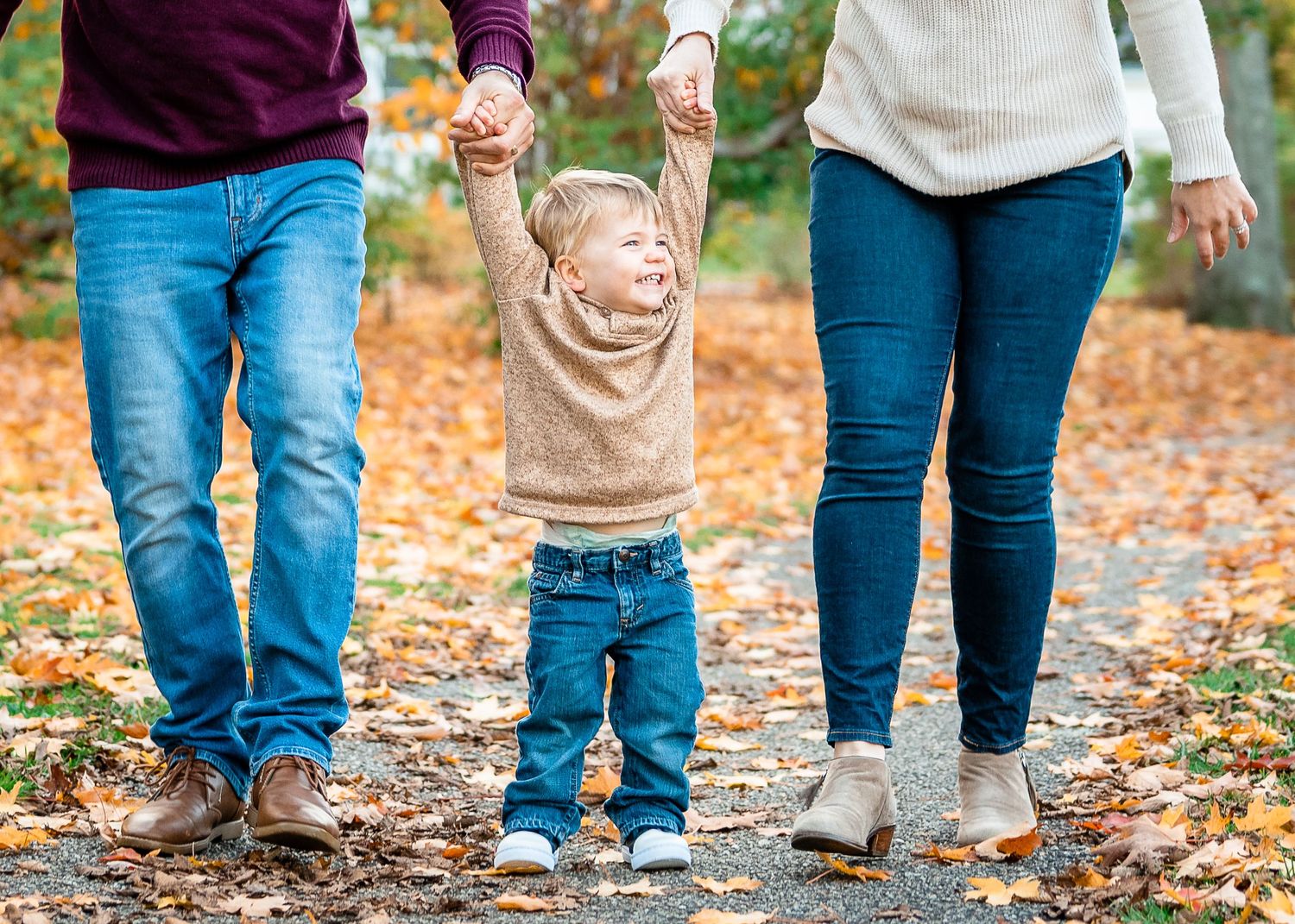 A family holds hands while walking through fallen leaves on an autumn afternoon.