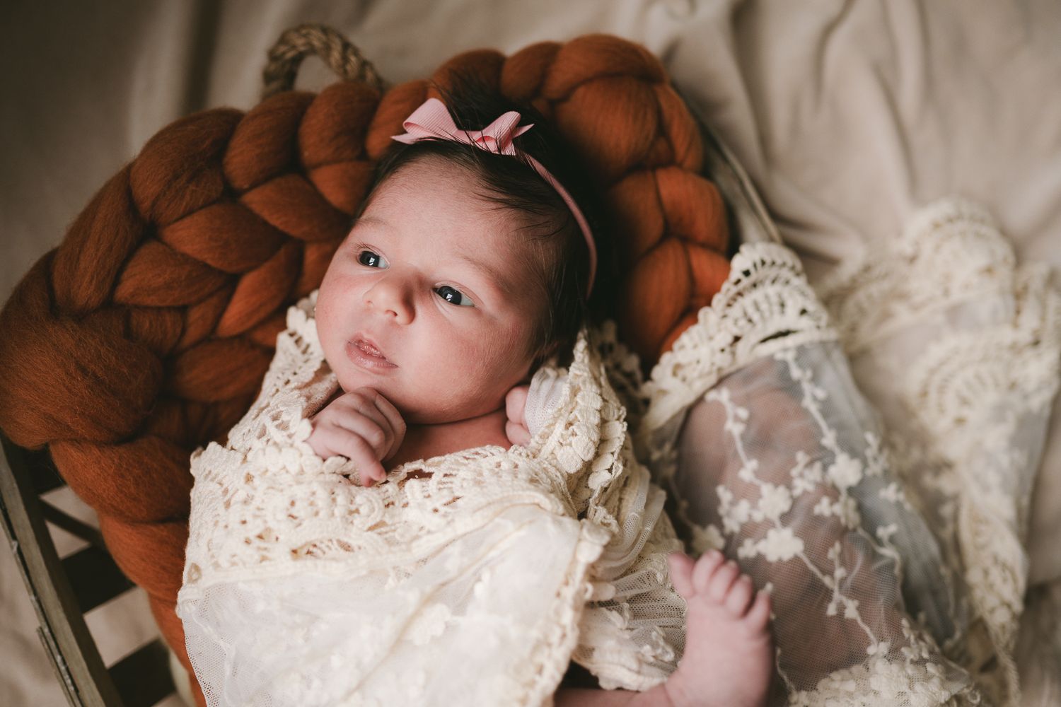 Newborn in delicate lace dress lies on a rustic brown knitted blanket.