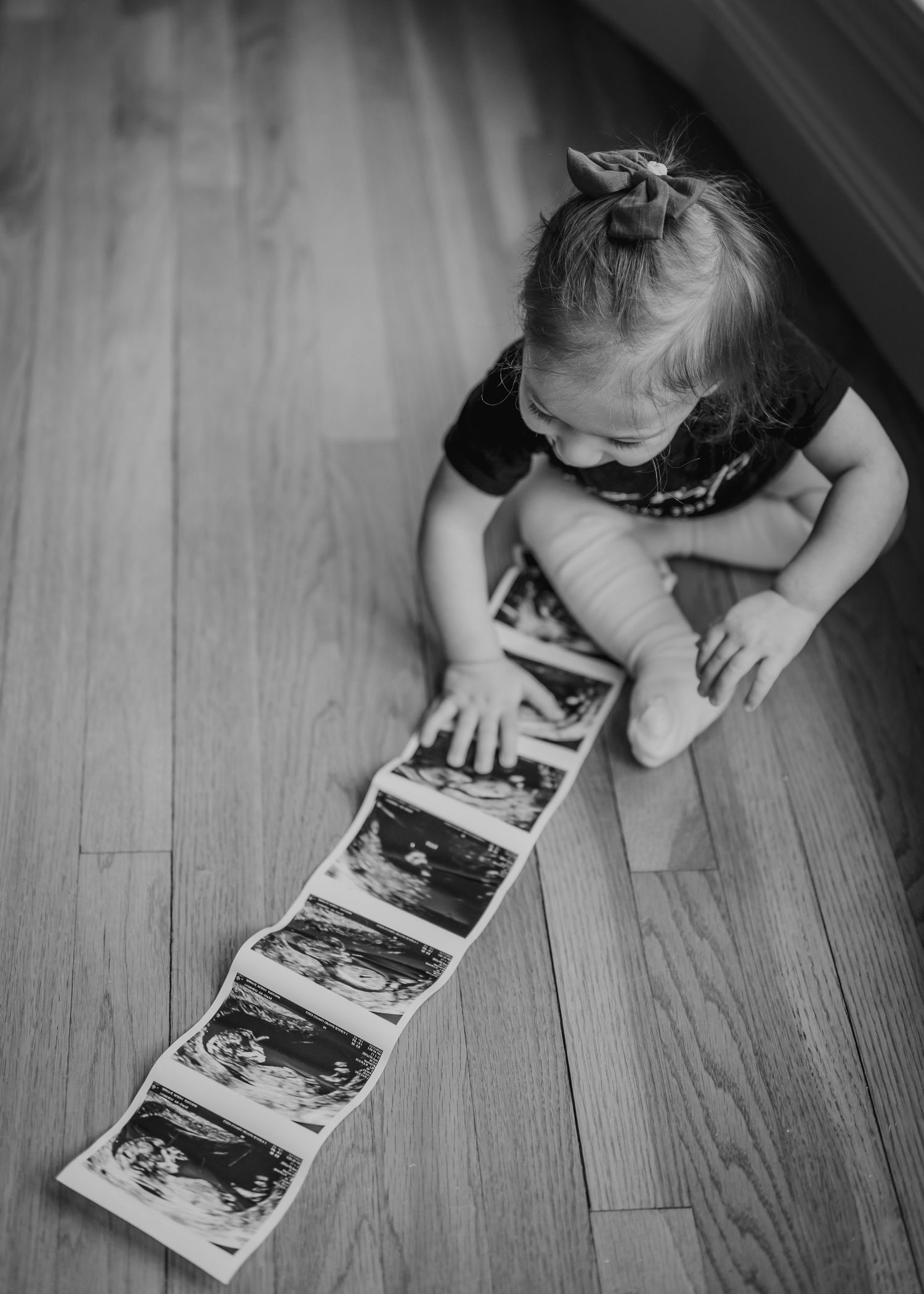 A toddler examines a strip of ultrasound photos on a hardwood floor.
