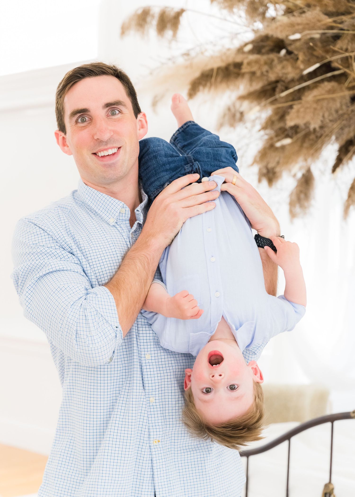 A parent holds a small child upside down playfully while smiling in a bright interior setting with decorative grasses.