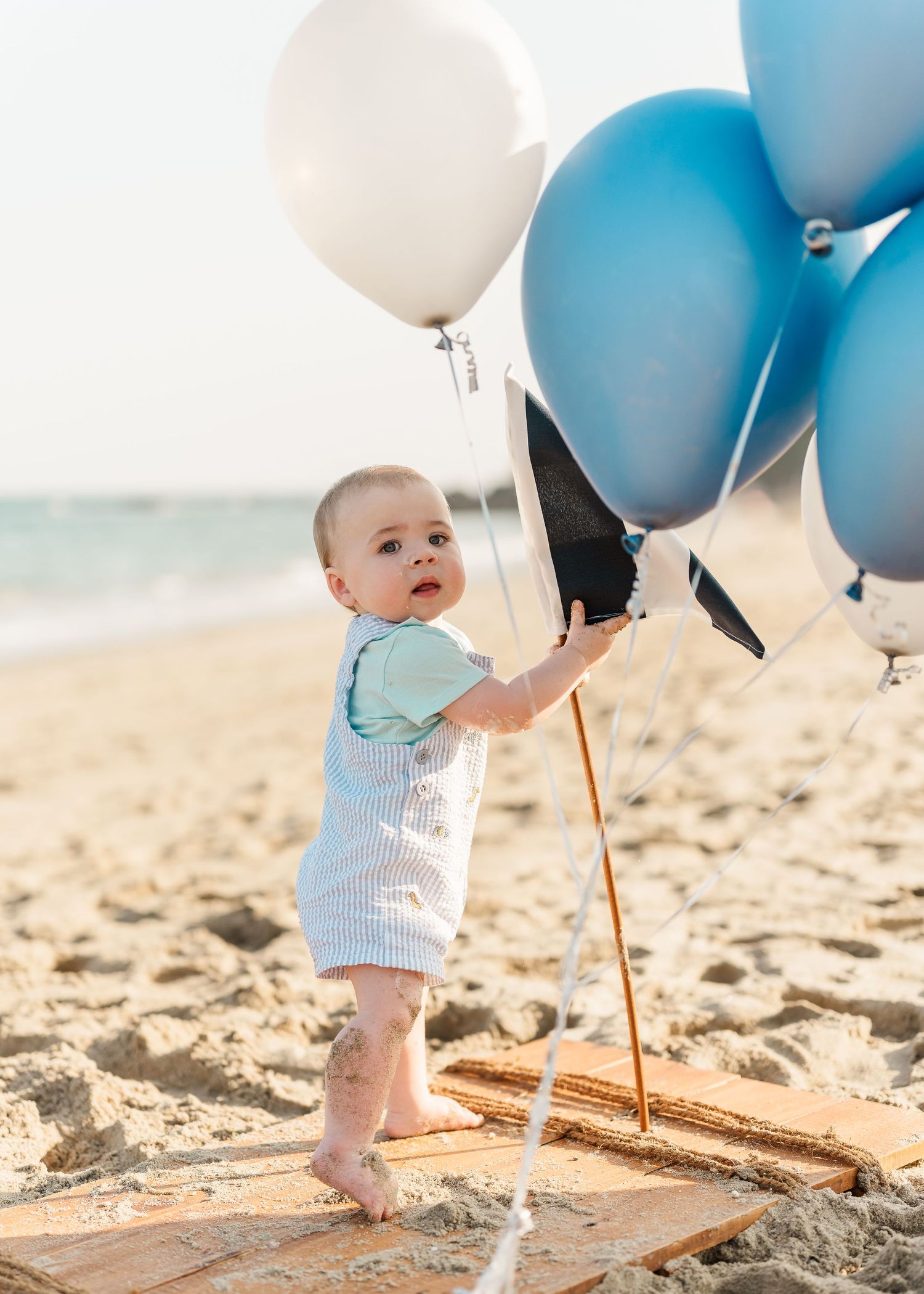 Baby's feet on sandy beach with light blue and white balloons creating a dreamy coastal celebration scene.