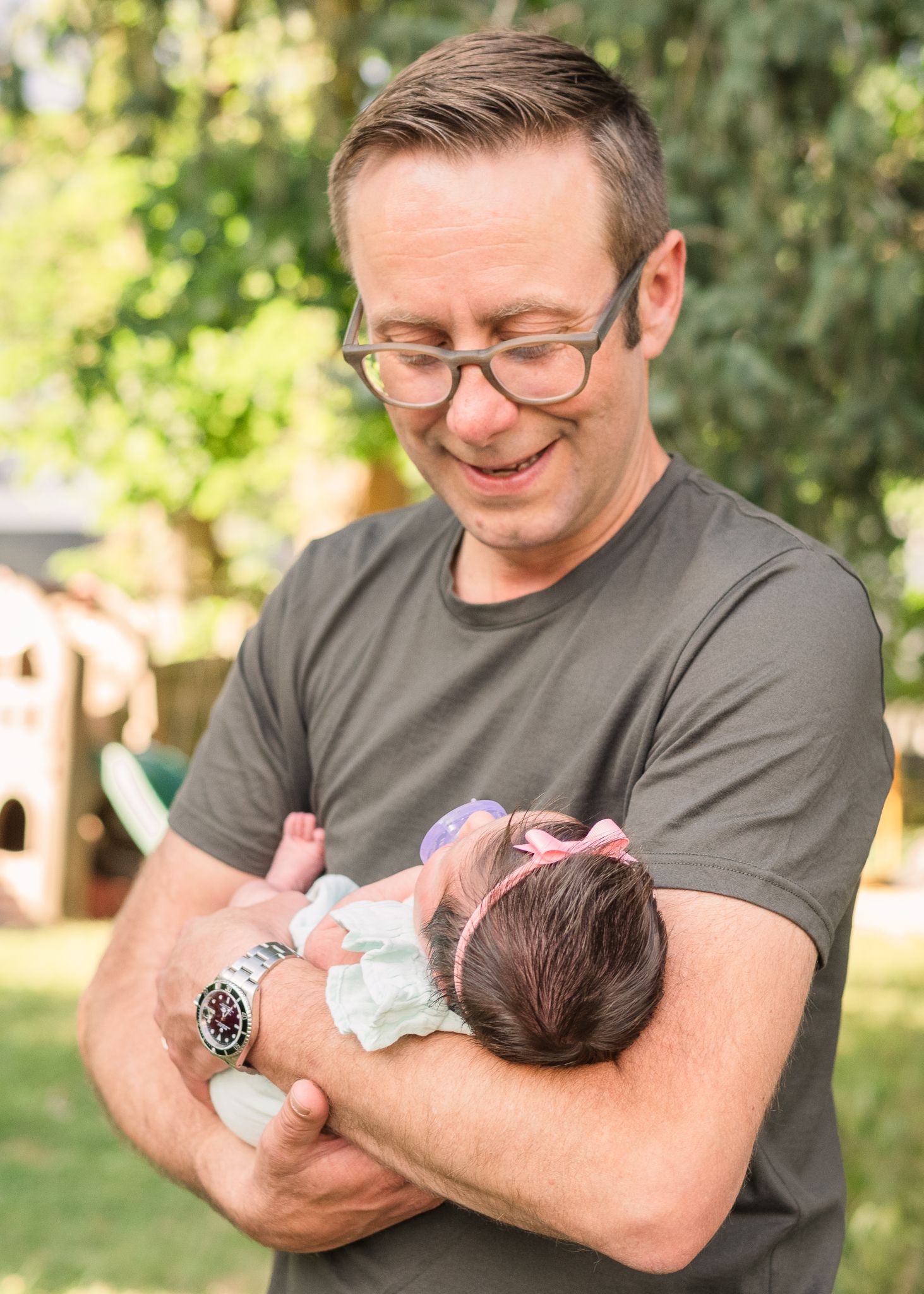 Adult in grey shirt tenderly holding newborn baby in sunlit garden setting.
