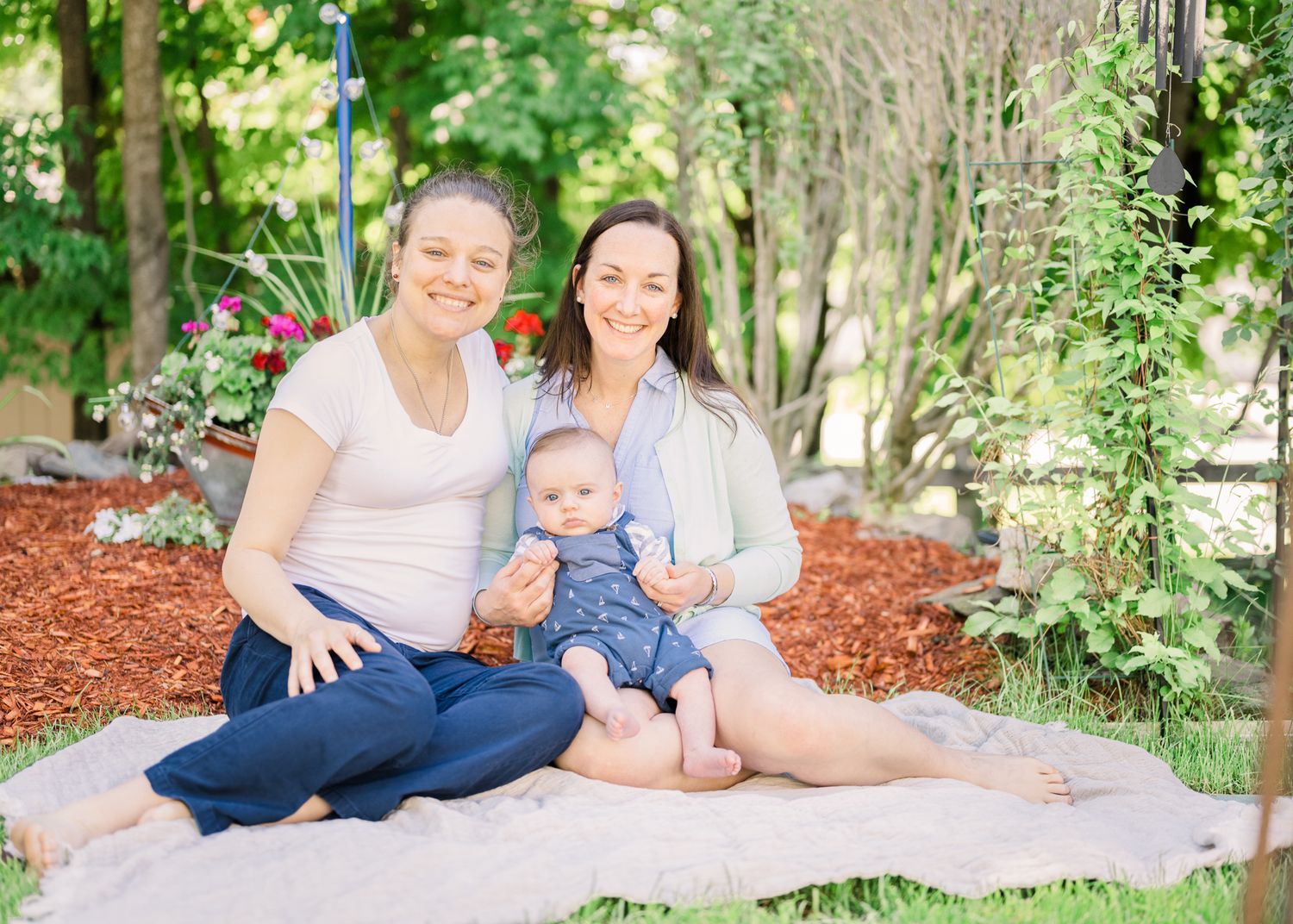 Family sits on blanket for portrait in lush garden setting with red mulch.