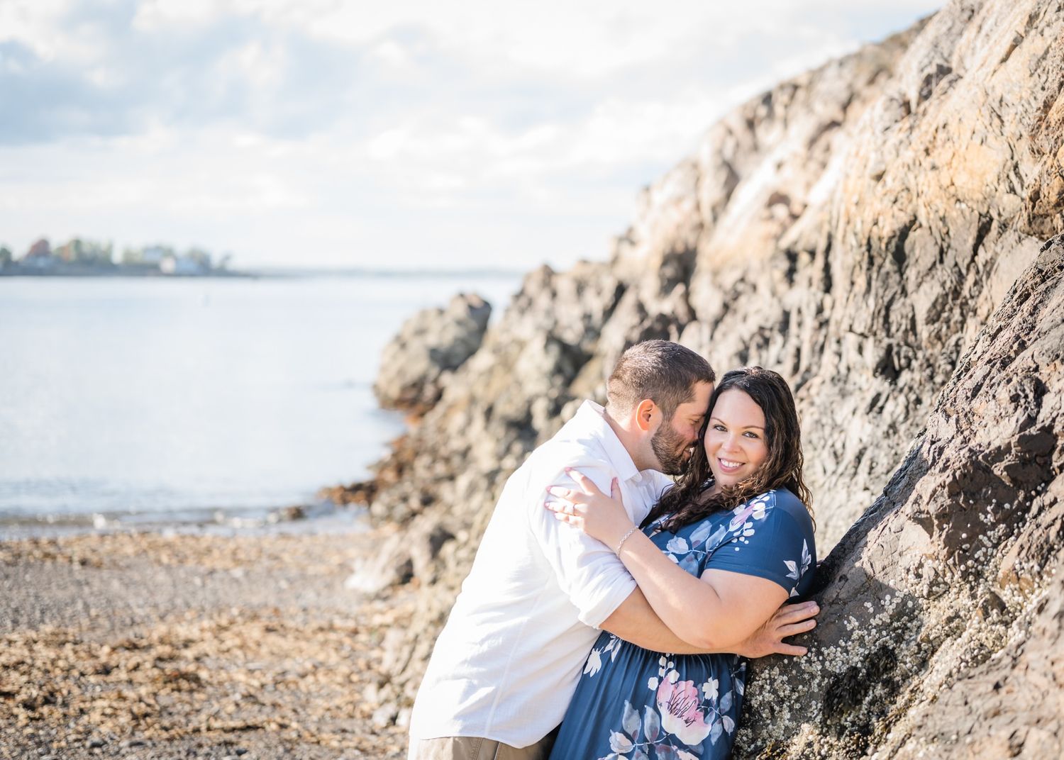 A couple embraces against a rocky coastal backdrop on a sunny day.