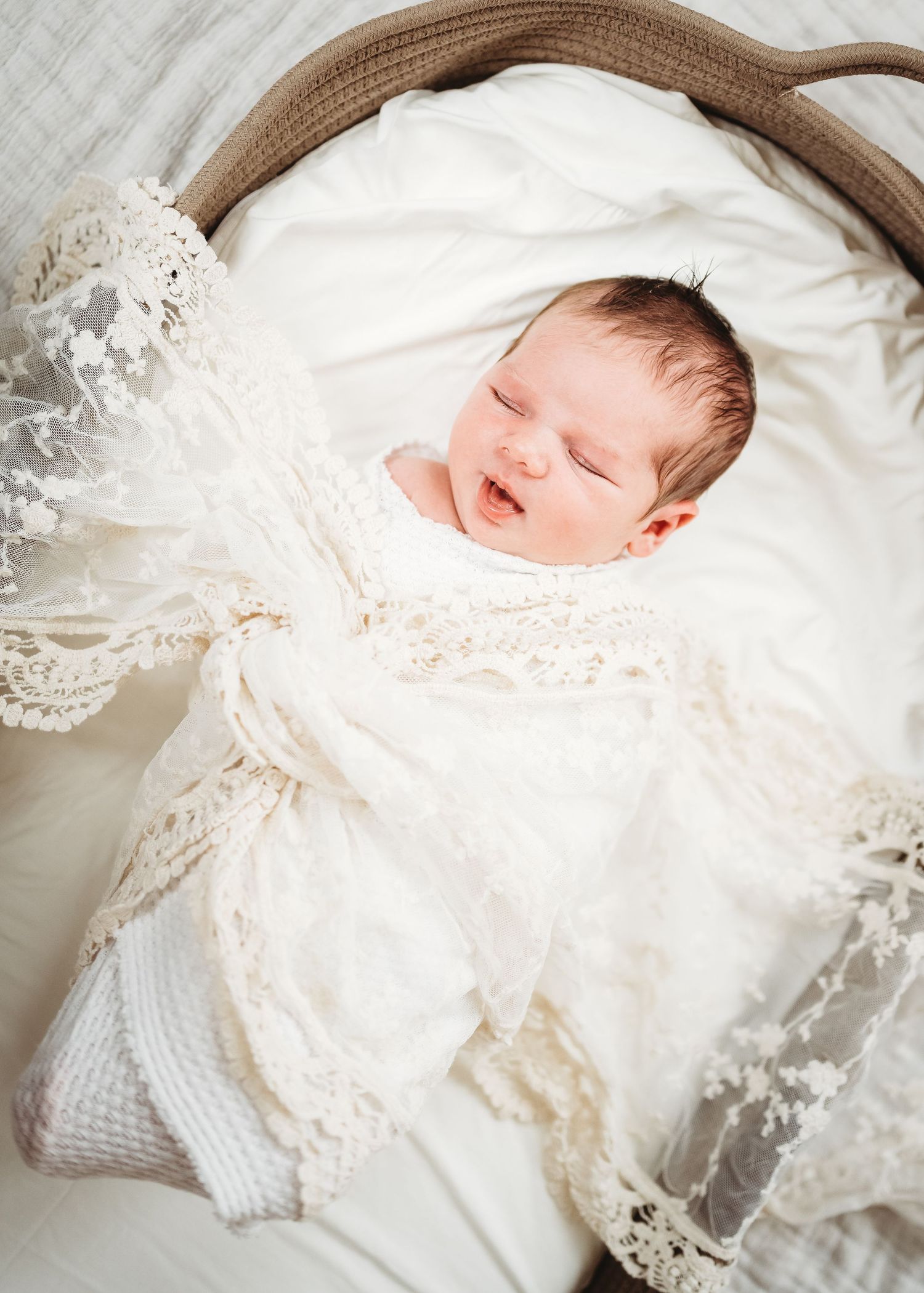 A sleeping newborn wearing delicate white lace and swaddled in soft white fabric rests peacefully on a textured surface.