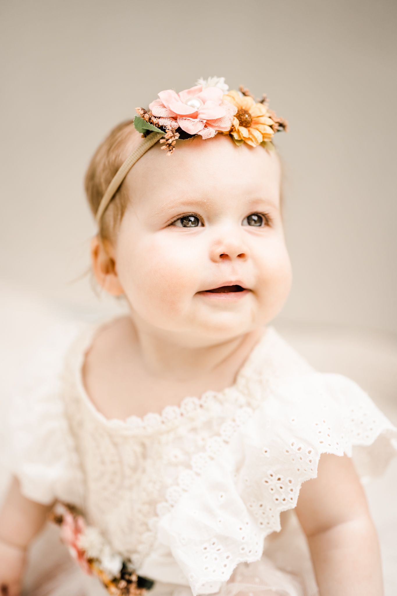 Baby wearing white eyelet dress and floral headband smiles while looking up.