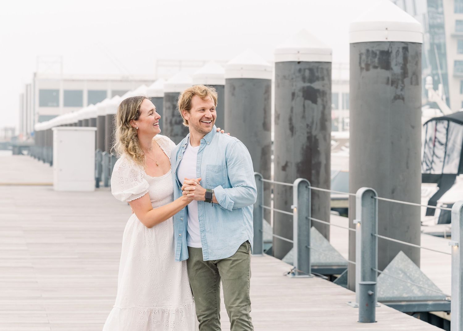 Couple walking along waterfront pier with industrial bollards and misty harbor views.