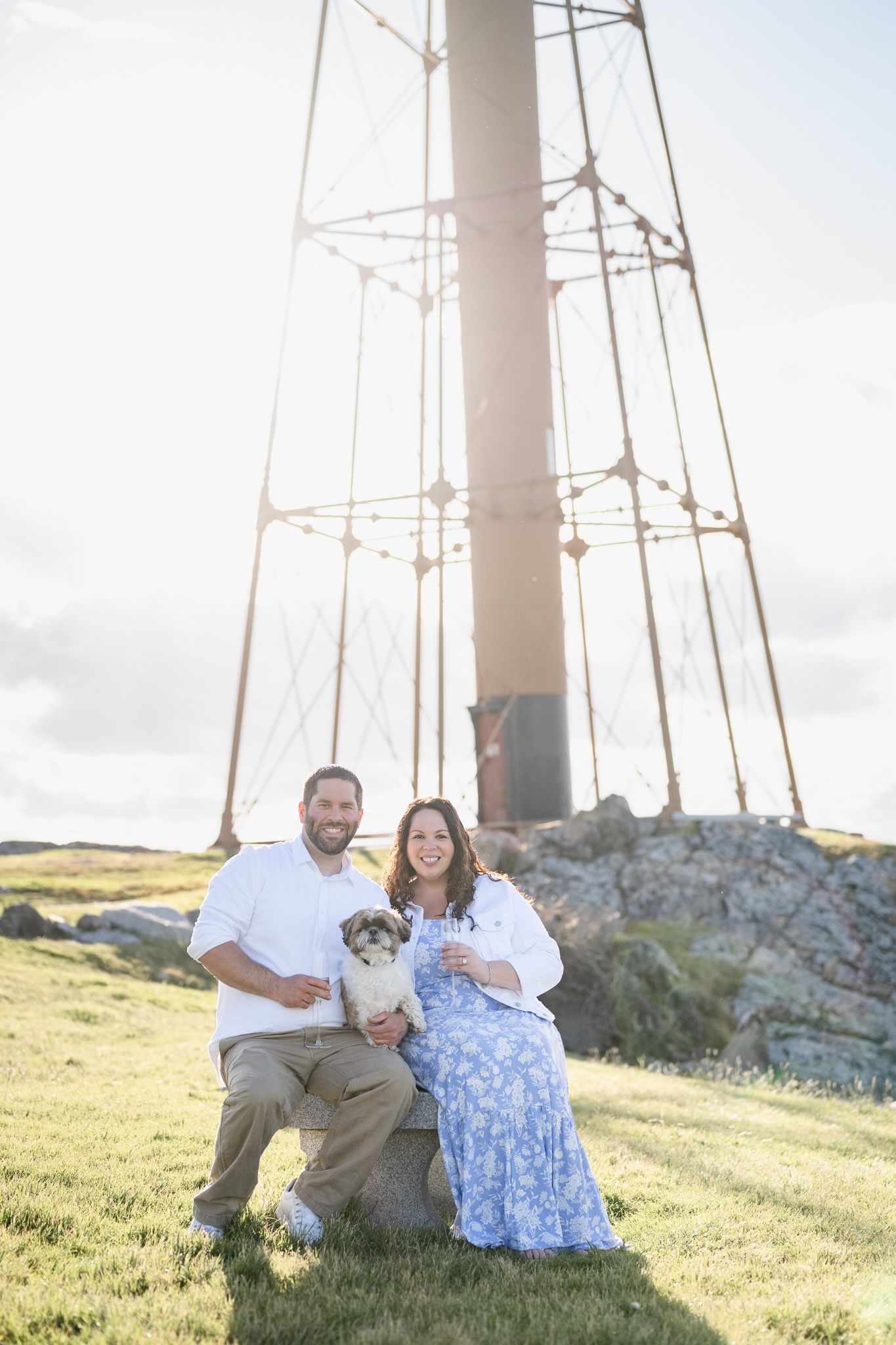 A couple and their dog pose together at the base of a lighthouse on a grassy hill.