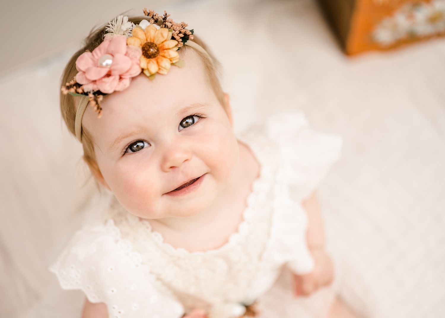 Baby wearing white dress and floral headband smiling at camera.