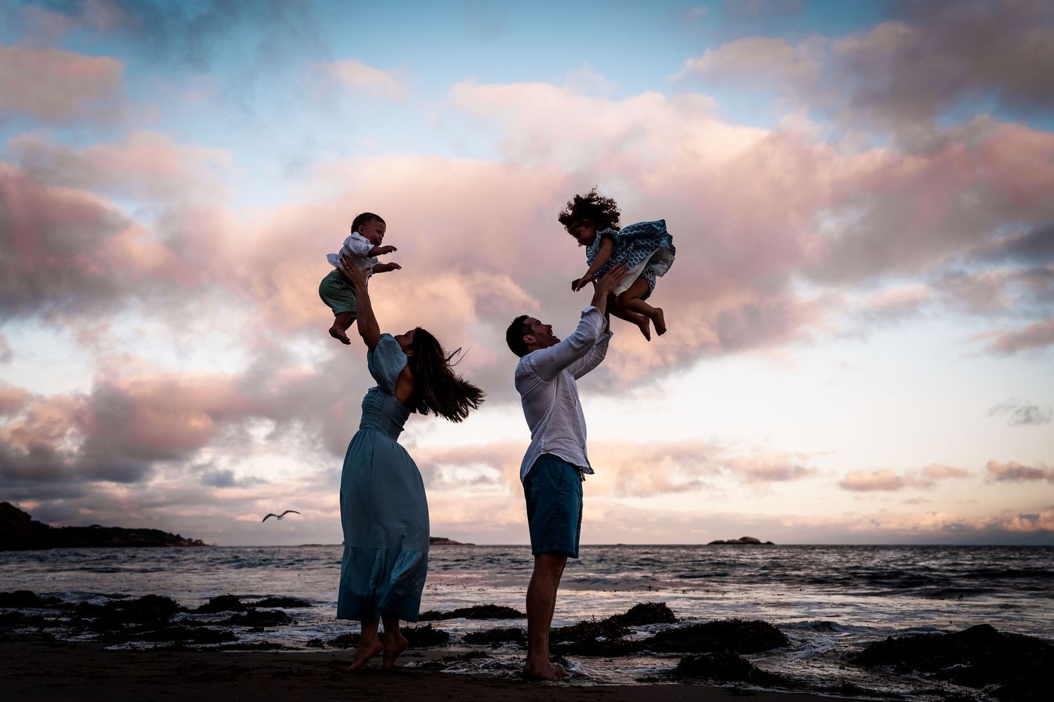 Silhouetted figures play with children against dramatic sunset sky at rocky beach.