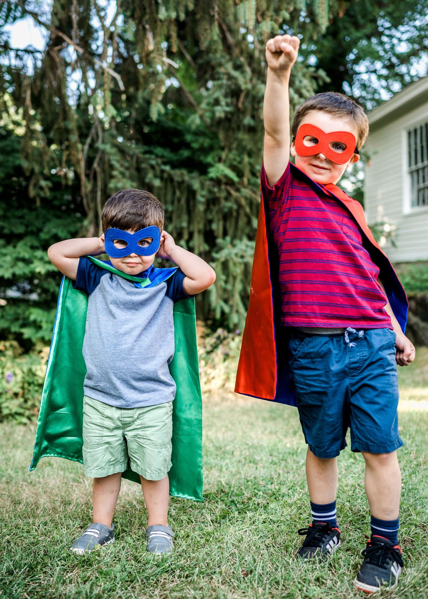 Two children wearing superhero masks and capes pose playfully in backyard.