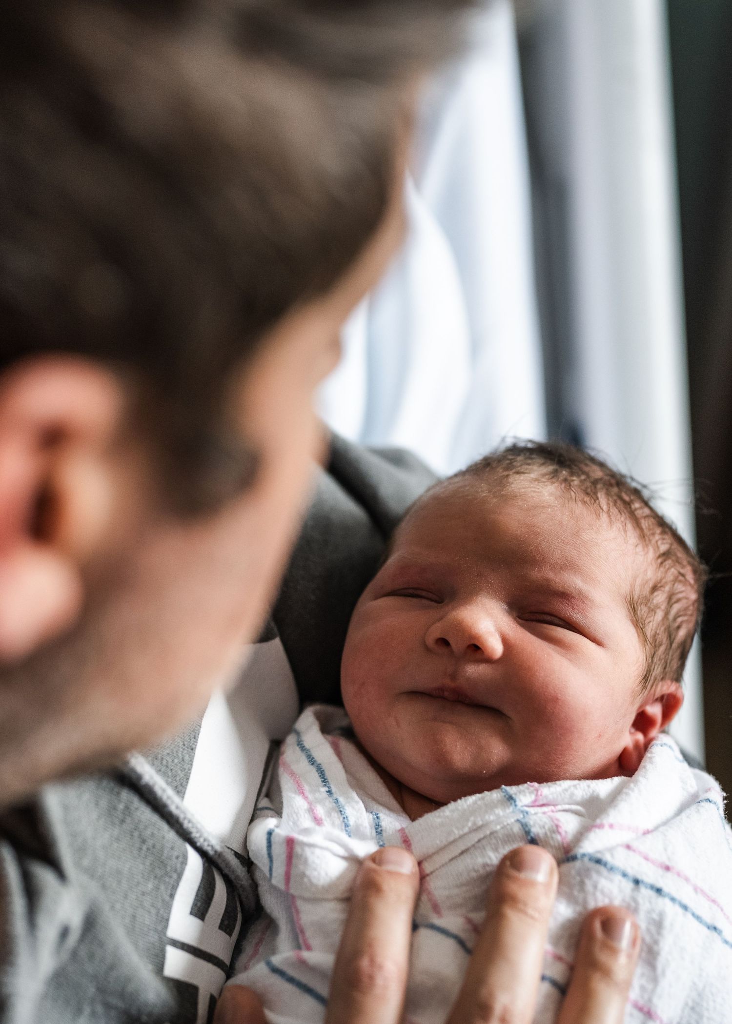 Newborn baby wrapped in white blanket smiles contentedly while being held near a window.
