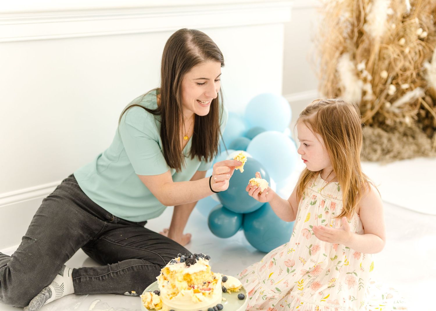 Woman in mint green shirt plays with toddler on floor with plush toy and yellow flower decorations.