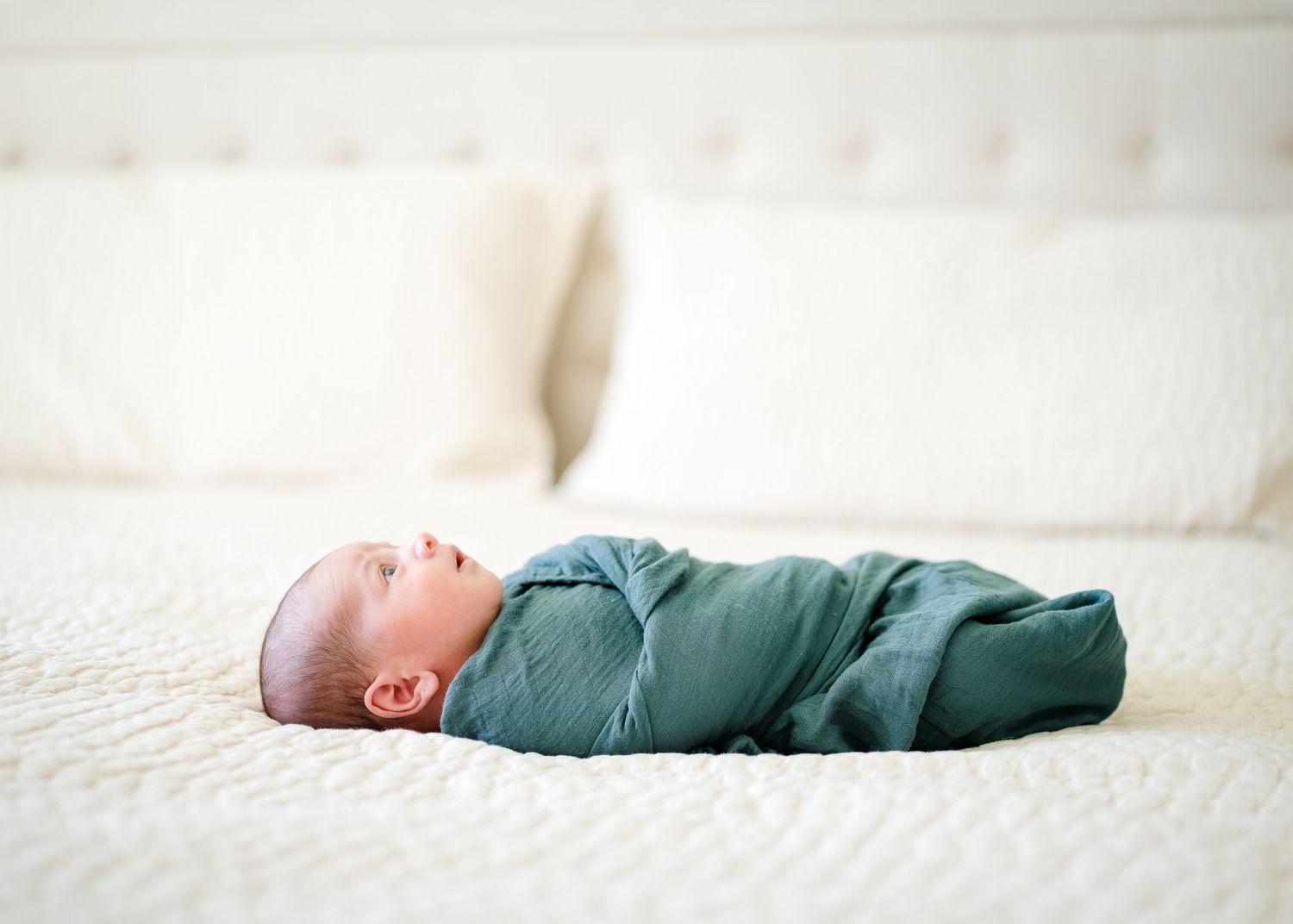 Sleeping baby wearing teal swaddle blanket lies on white bedding.