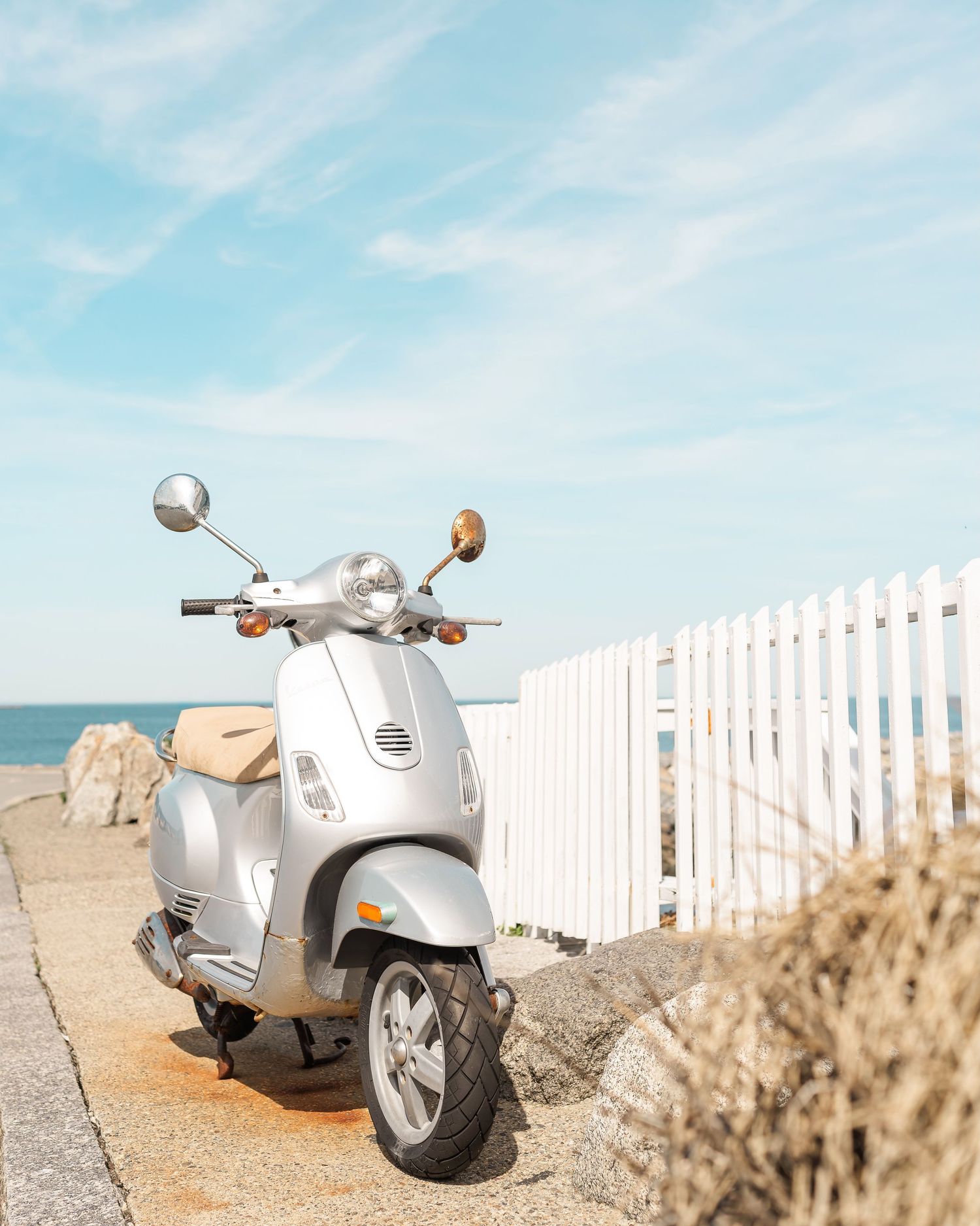 White vintage Vespa scooter parked near white picket fence on sandy beach with ocean view in background.