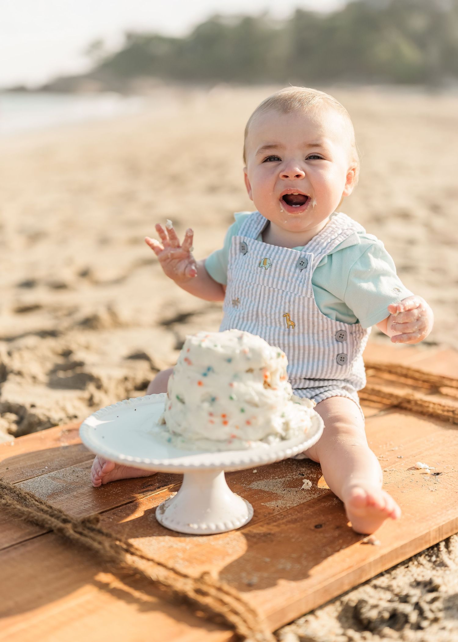 Baby in striped outfit sits on wooden surface at beach with small cake and joyfully plays with frosting.