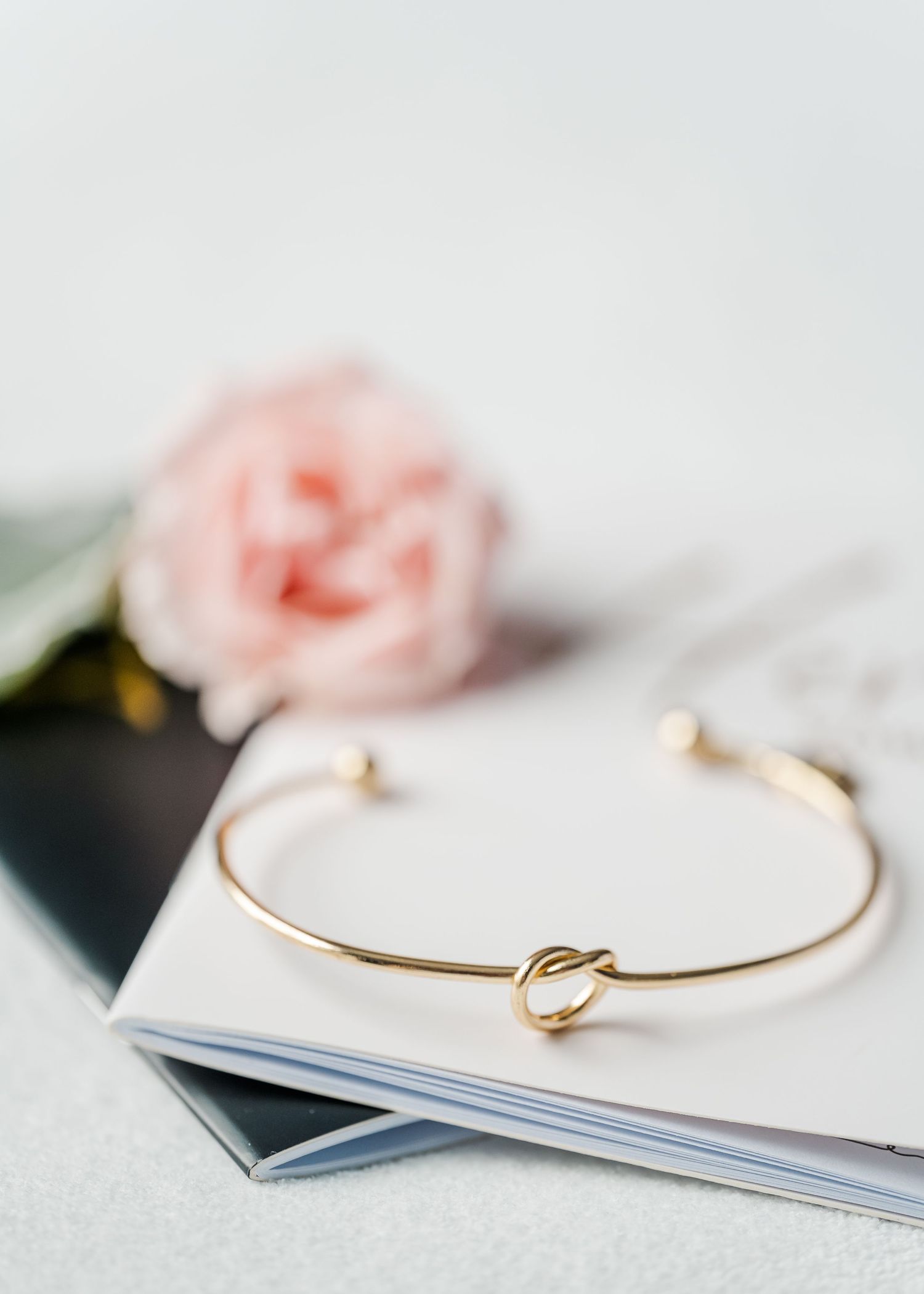 Delicate gold knot bangle bracelet displayed on white surface with soft pink flowers in background.