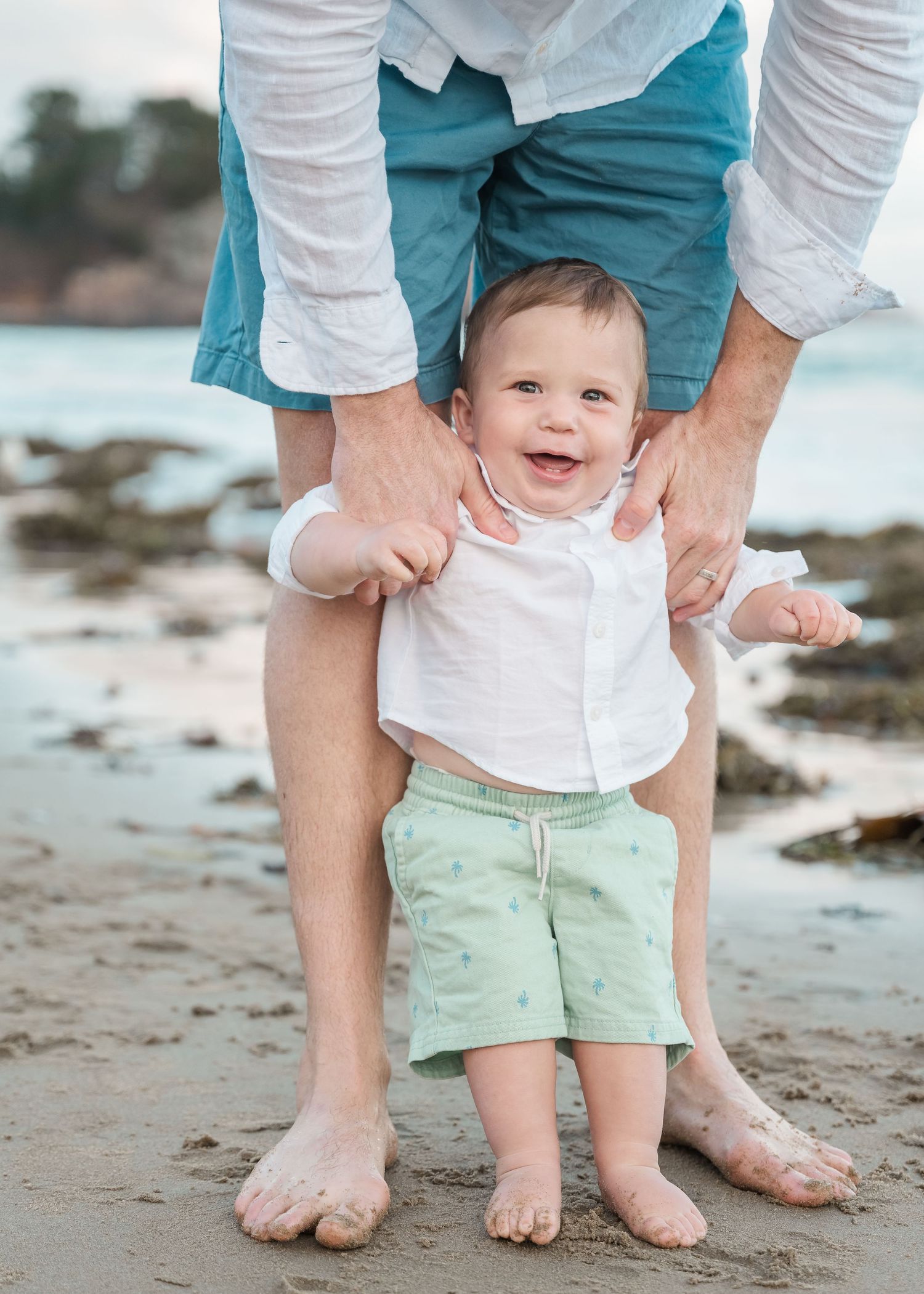 Small child in white shirt and mint shorts learns to walk on beach with parent's support while showing happy expression.