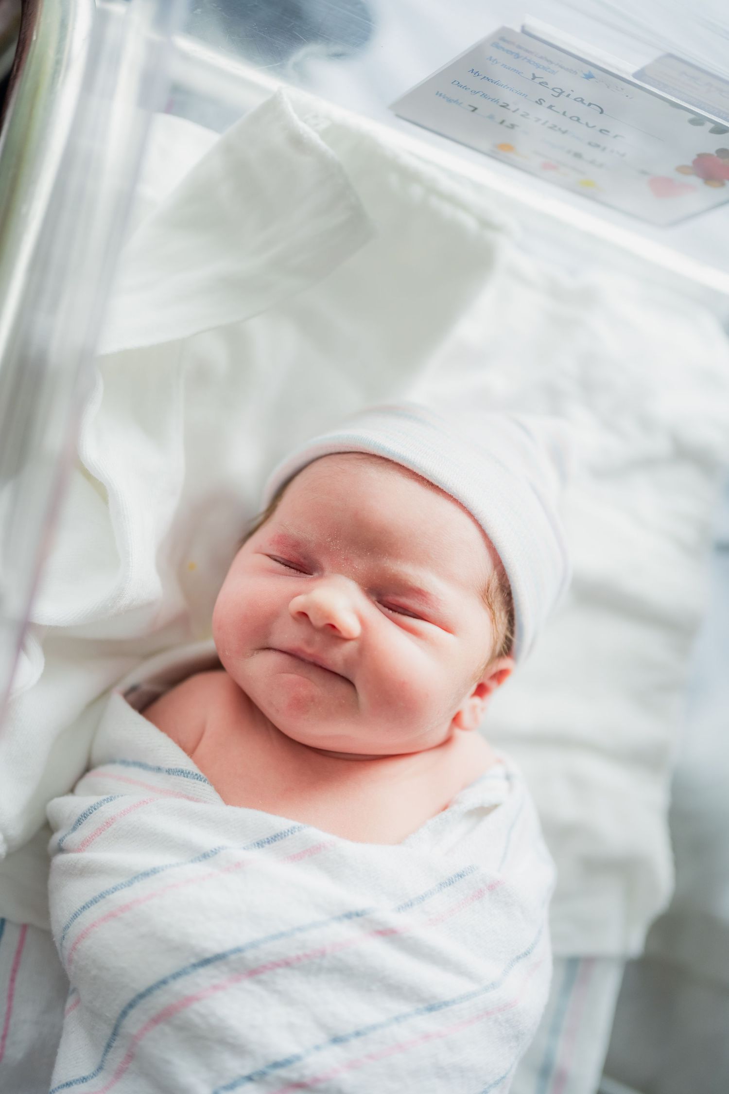 A newborn baby wrapped in a striped blanket smiles while resting in a hospital bassinet.