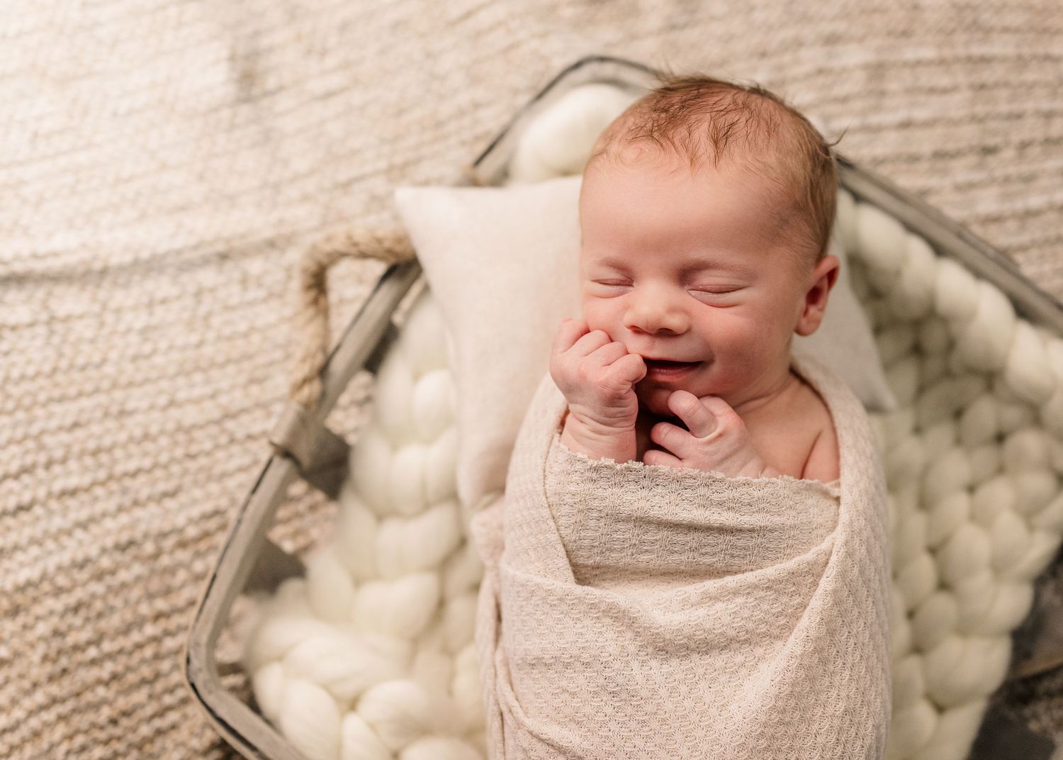 A newborn baby yawns while swaddled in a beige wrap, lying on a textured backdrop with cozy white pom poms.