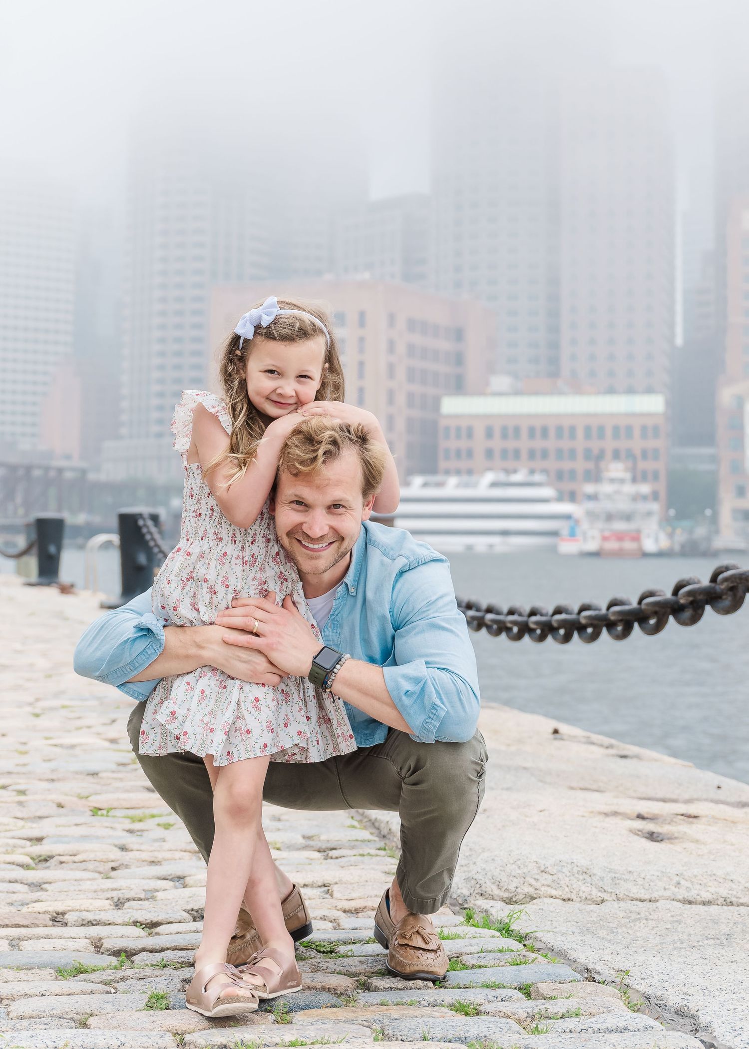 A heartwarming moment between father and daughter on a waterfront pier with foggy city skyline.