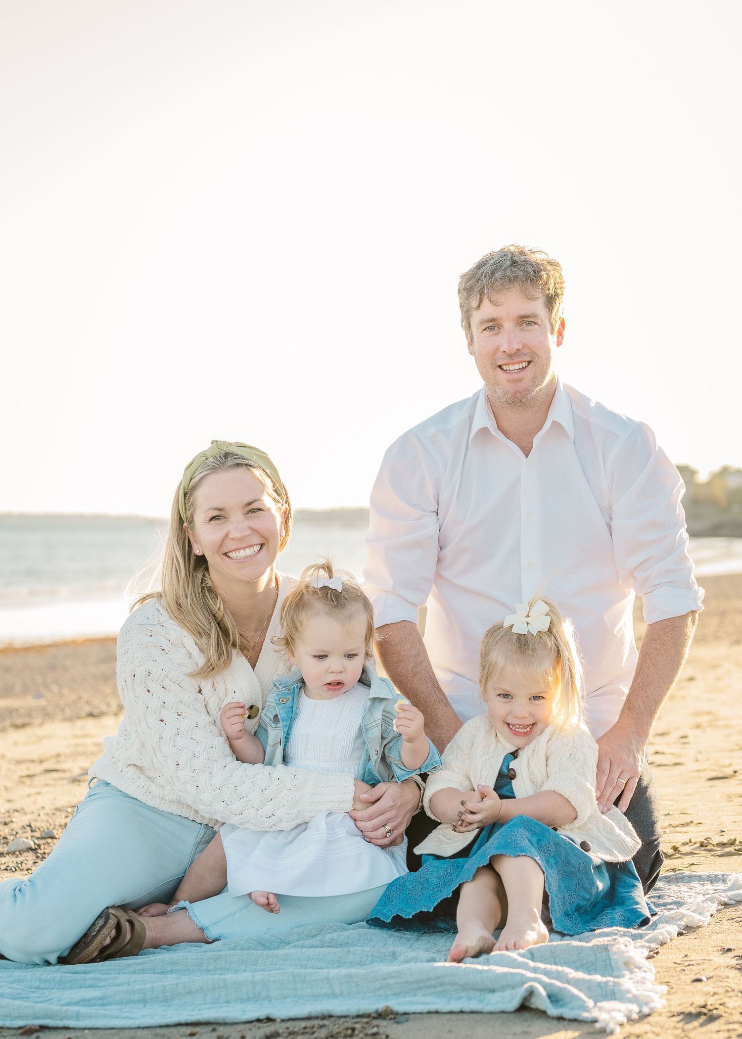 Family sitting together on blue blanket at beach during golden hour photo session.
