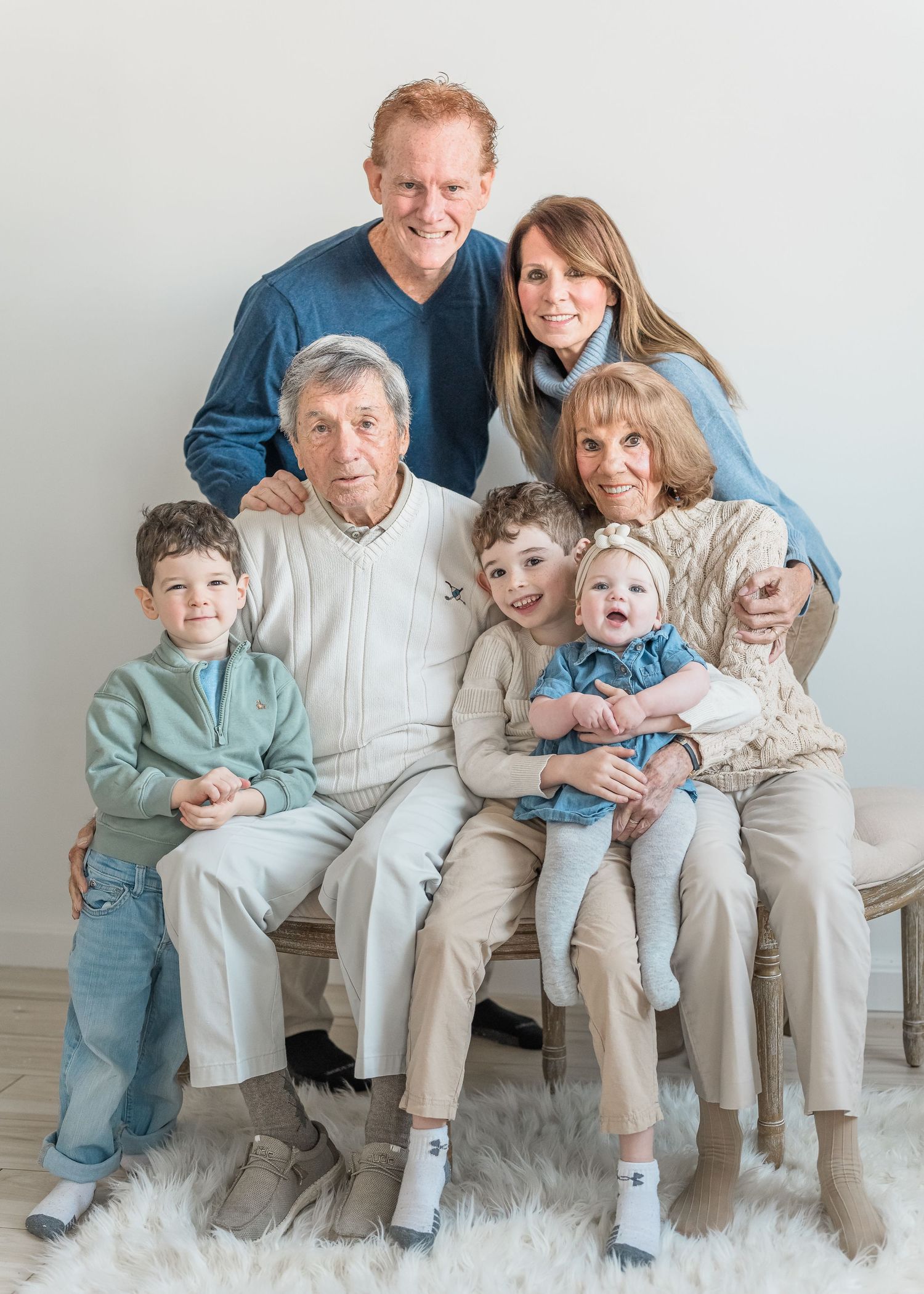 A family wearing coordinated blue and beige outfits sits together on a white couch for a portrait session.