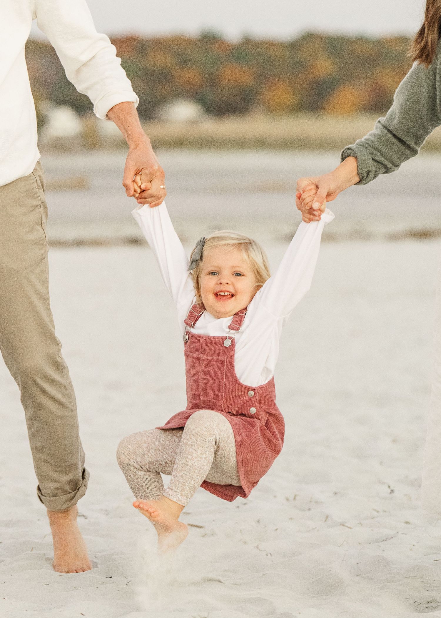 Child in red overalls being swung playfully by adults on a beach, creating joyful moments.