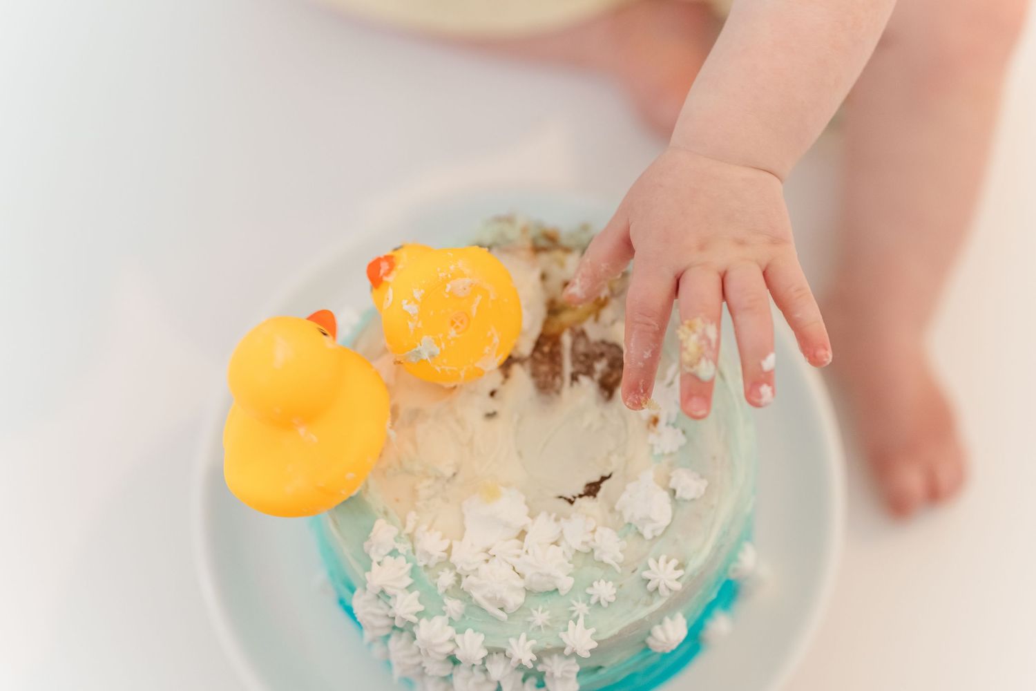 Cake smash. A yellow rubber duck toy floats in a bubble-filled sink as hands play with the soapy water.