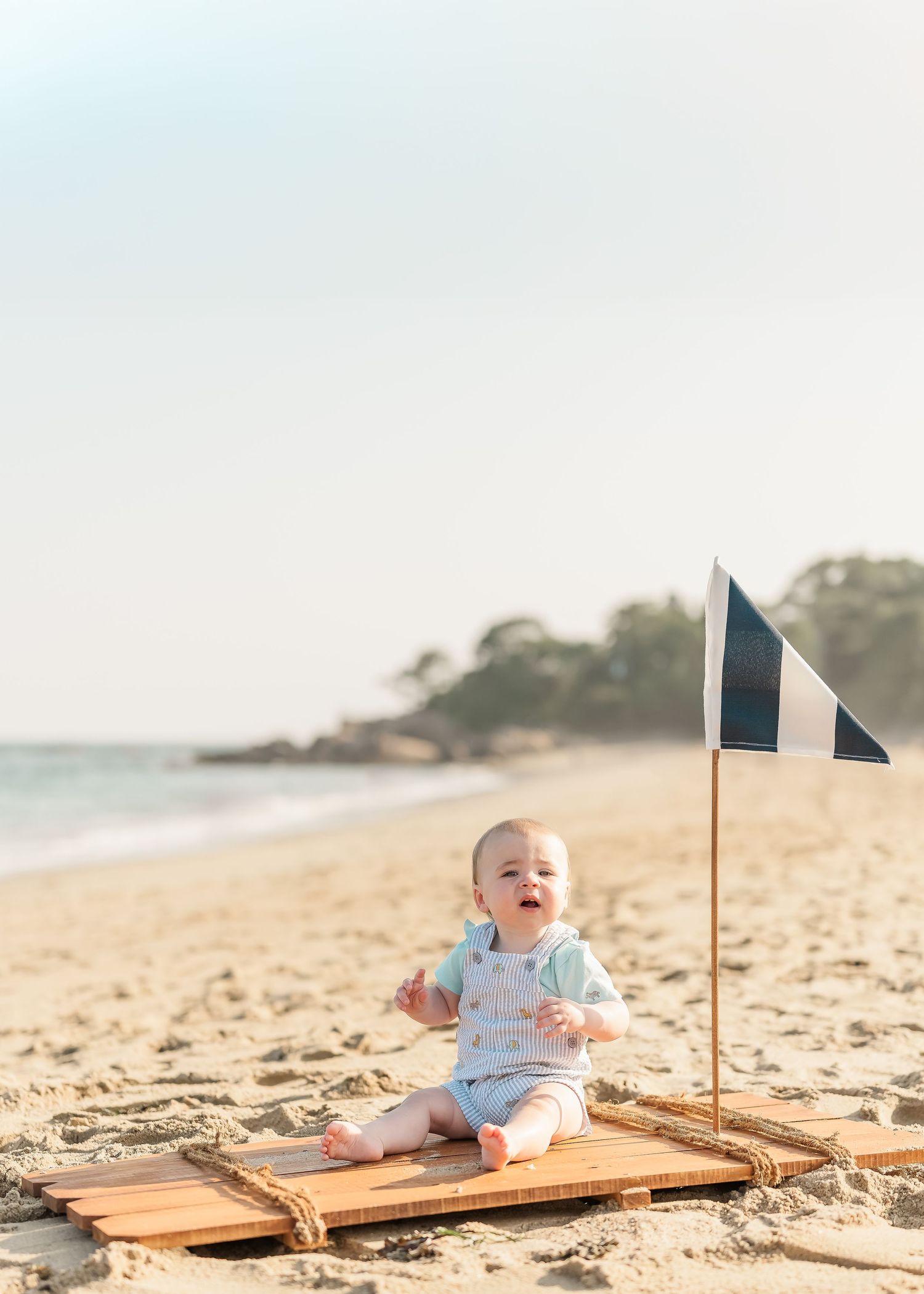 Baby sits on wooden raft with small flags on sandy beach with ocean waves in background.