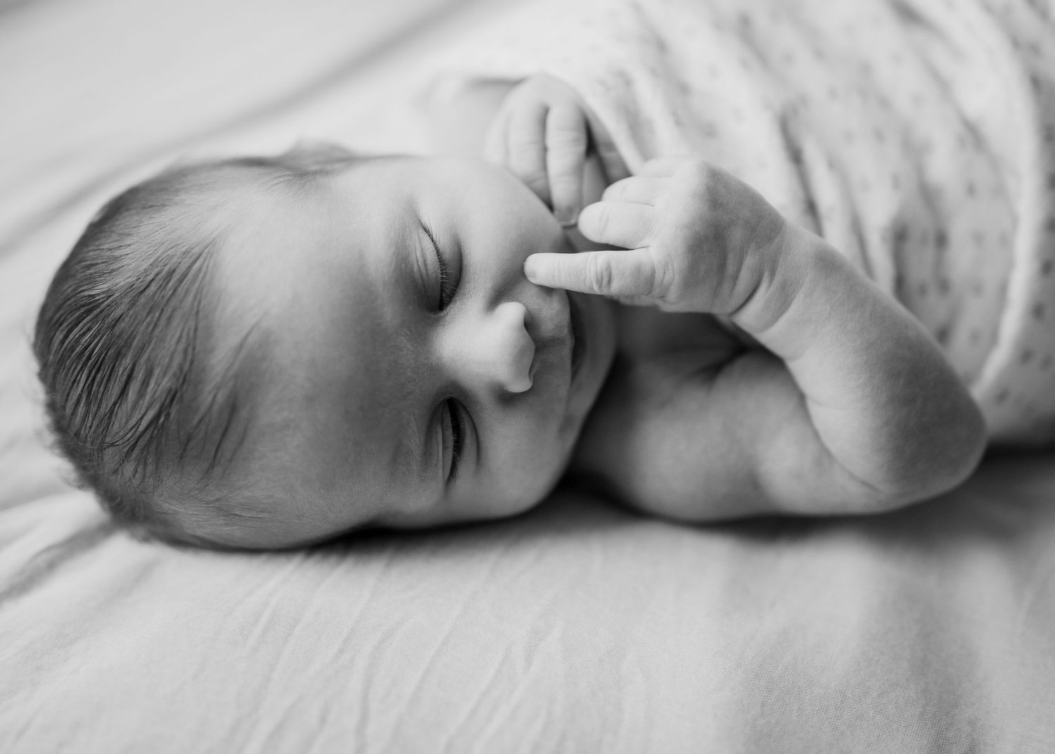 Black and white closeup of sleeping baby with hand near face on white bedding.