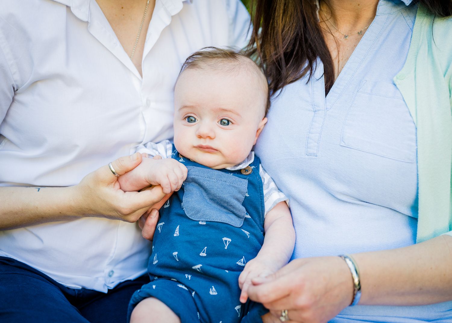 A small baby in a blue outfit is held lovingly between two people.