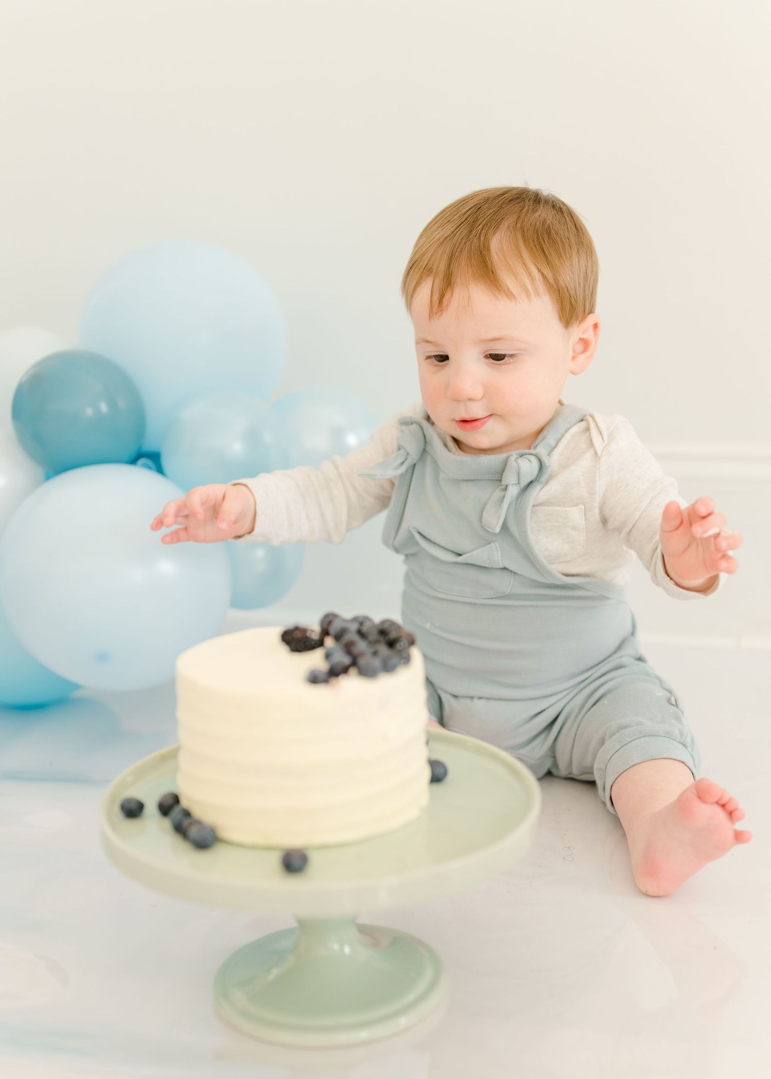 A minimalist white cake with blueberries sits on a mint green cake stand with soft blue balloons in the background.