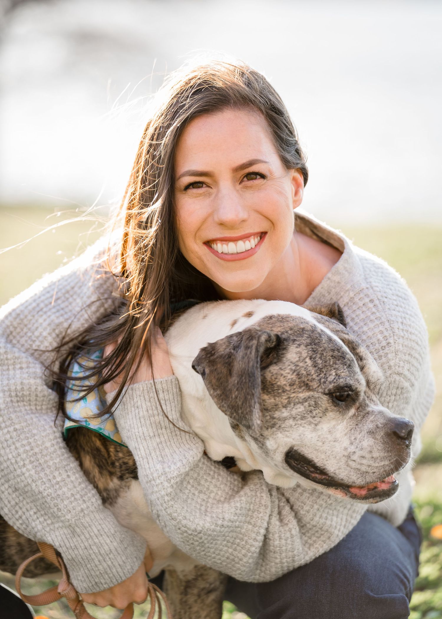 A person in a gray sweater hugs and smiles with their senior gray pit bull dog outdoors.