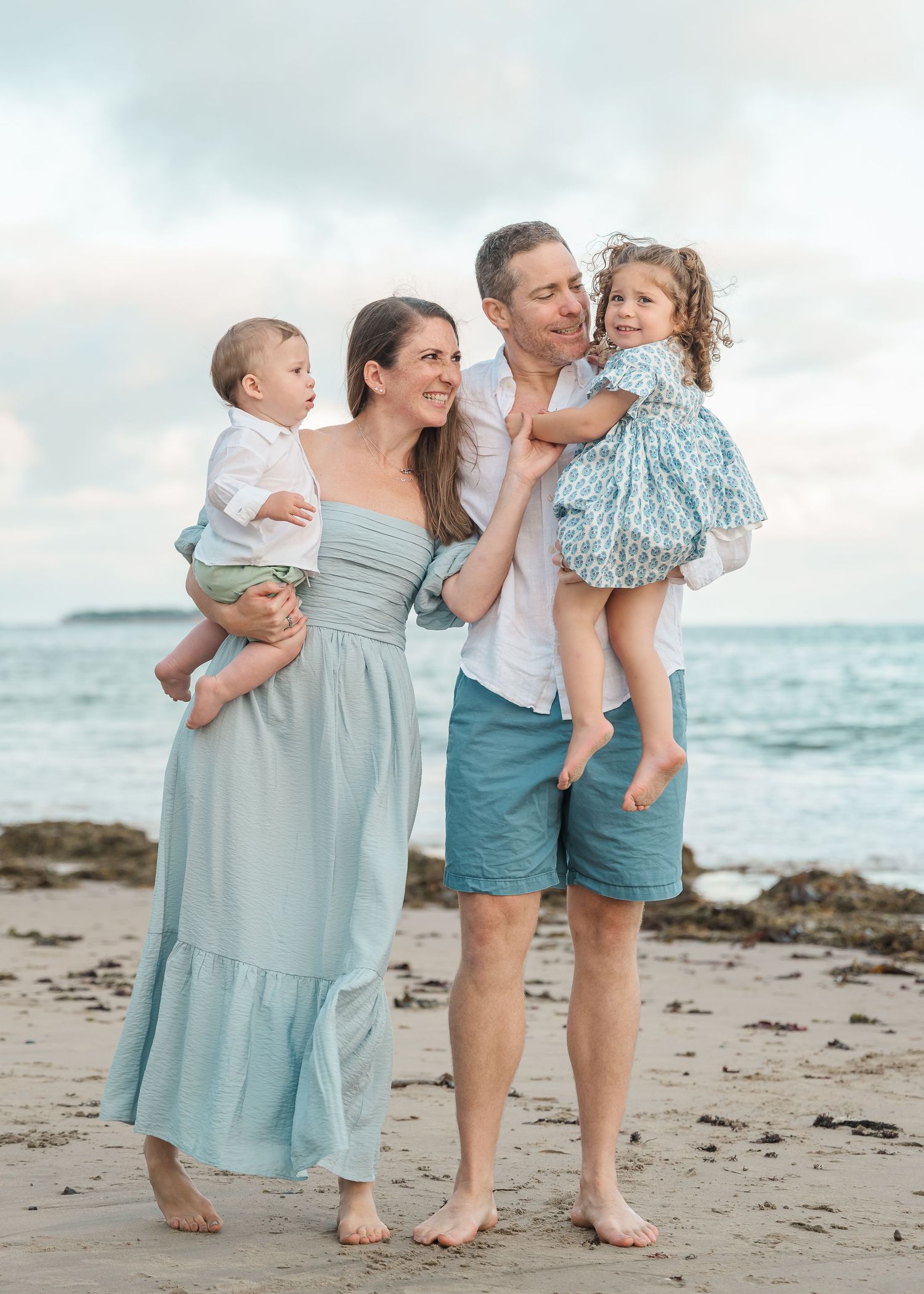 Family in coordinating blue outfits stands together on beach at sunset with ocean and rocks in background.