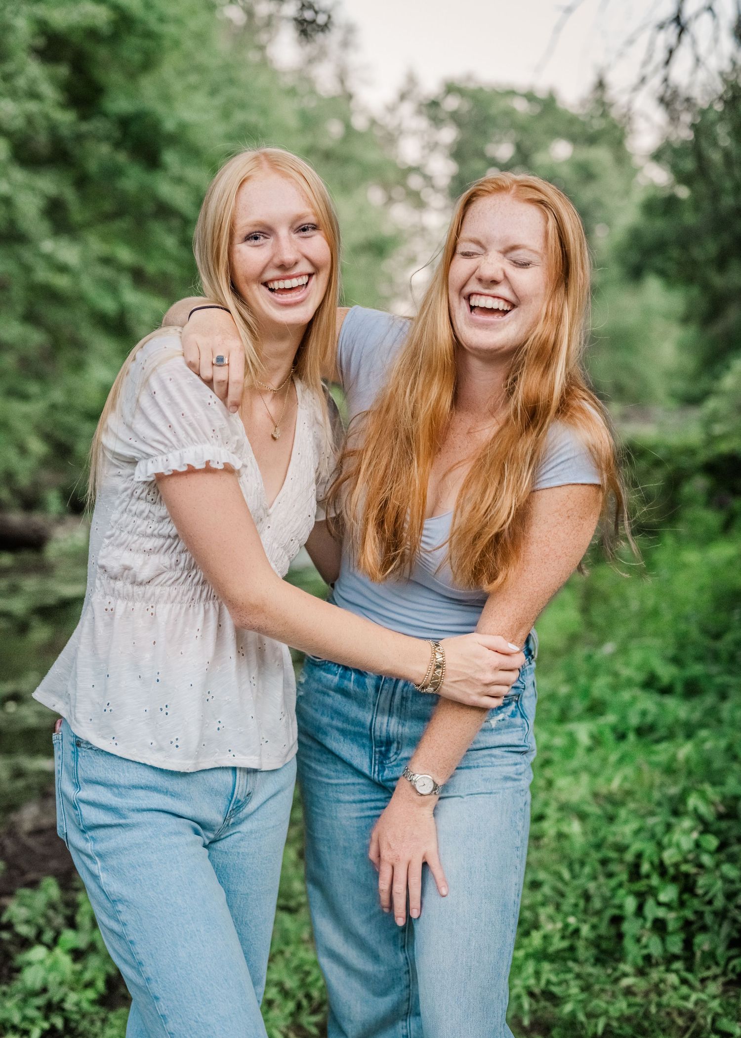 Two people share a candid laughing moment while hugging in casual outfits against green foliage.